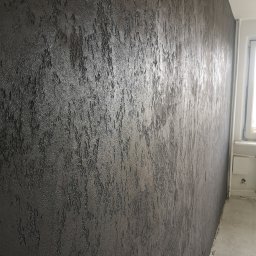 LEOPOL SPÓŁKA Z OGRANICZONĄ ODPOWIEDZIALNOŚCIĄ - Ciemna ściana z wyraźną, nieregularną fakturą, przypominającą korę drzewa, widoczna z boku, z fragmentem okna w tle.