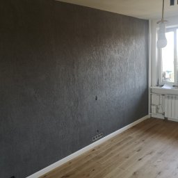 LEOPOL SPÓŁKA Z OGRANICZONĄ ODPOWIEDZIALNOŚCIĄ - Ściana z dekoracyjną, ciemną fakturą w mieszkaniu w Warszawie, widoczne gniazdka elektryczne i fragment podłogi z jasnego drewna oraz biały grzejnik pod oknem.