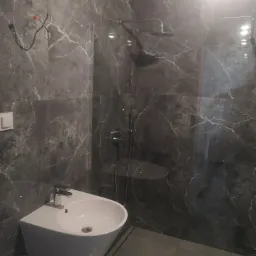 Wykończona łazienka z kabiną prysznicową typu walk-in, umywalką nablatową na postumencie i szarą, marmurkową okładziną ścienną z widocznymi przewodami elektrycznymi.