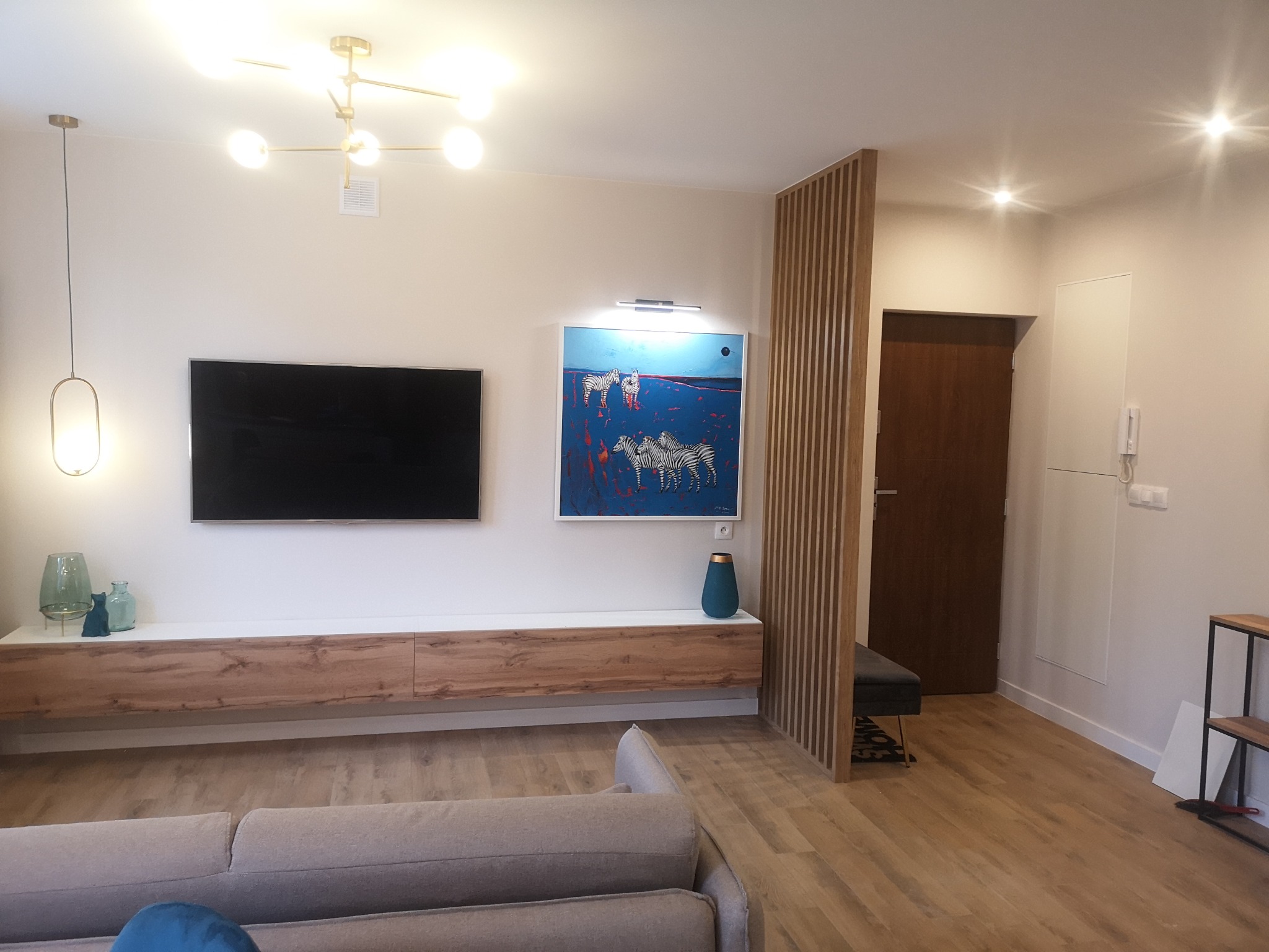 Jasne wnętrze salonu z drewnianą podłogą, telewizorem, szafką RTV z drewnianym frontem, obrazem z zebrami i drewnianą ścianką działową z pionowych listew.