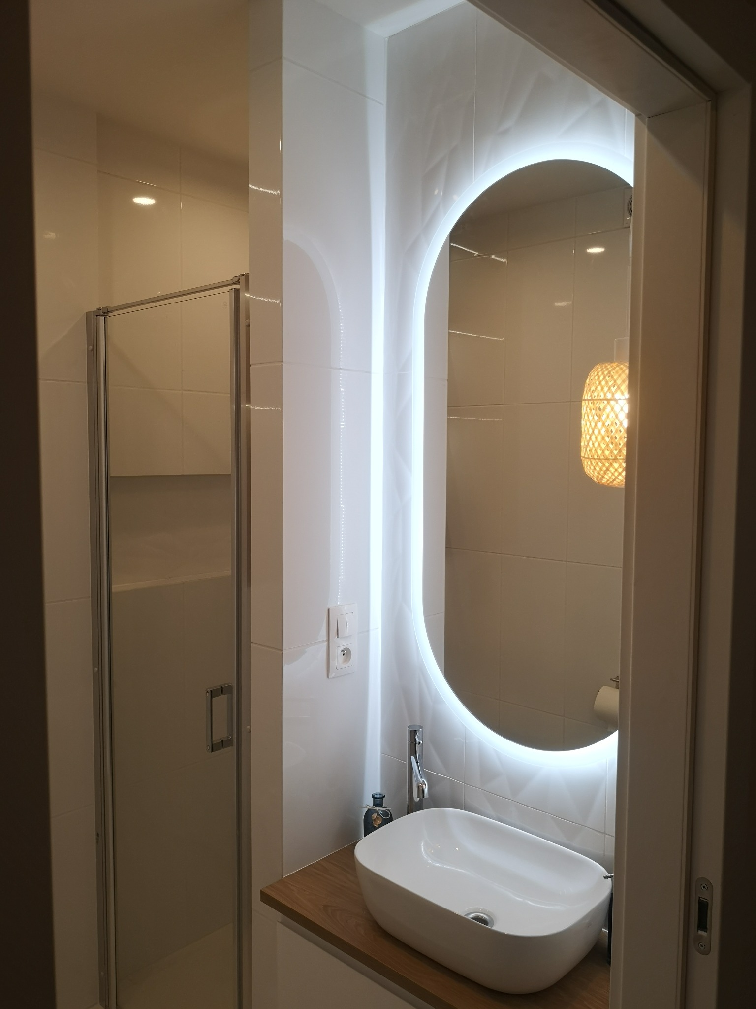 Nowoczesna łazienka z kabiną prysznicową, umywalką nablatową i owalnym lustrem z podświetleniem LED, wisząca lampa z wikliny w tle.