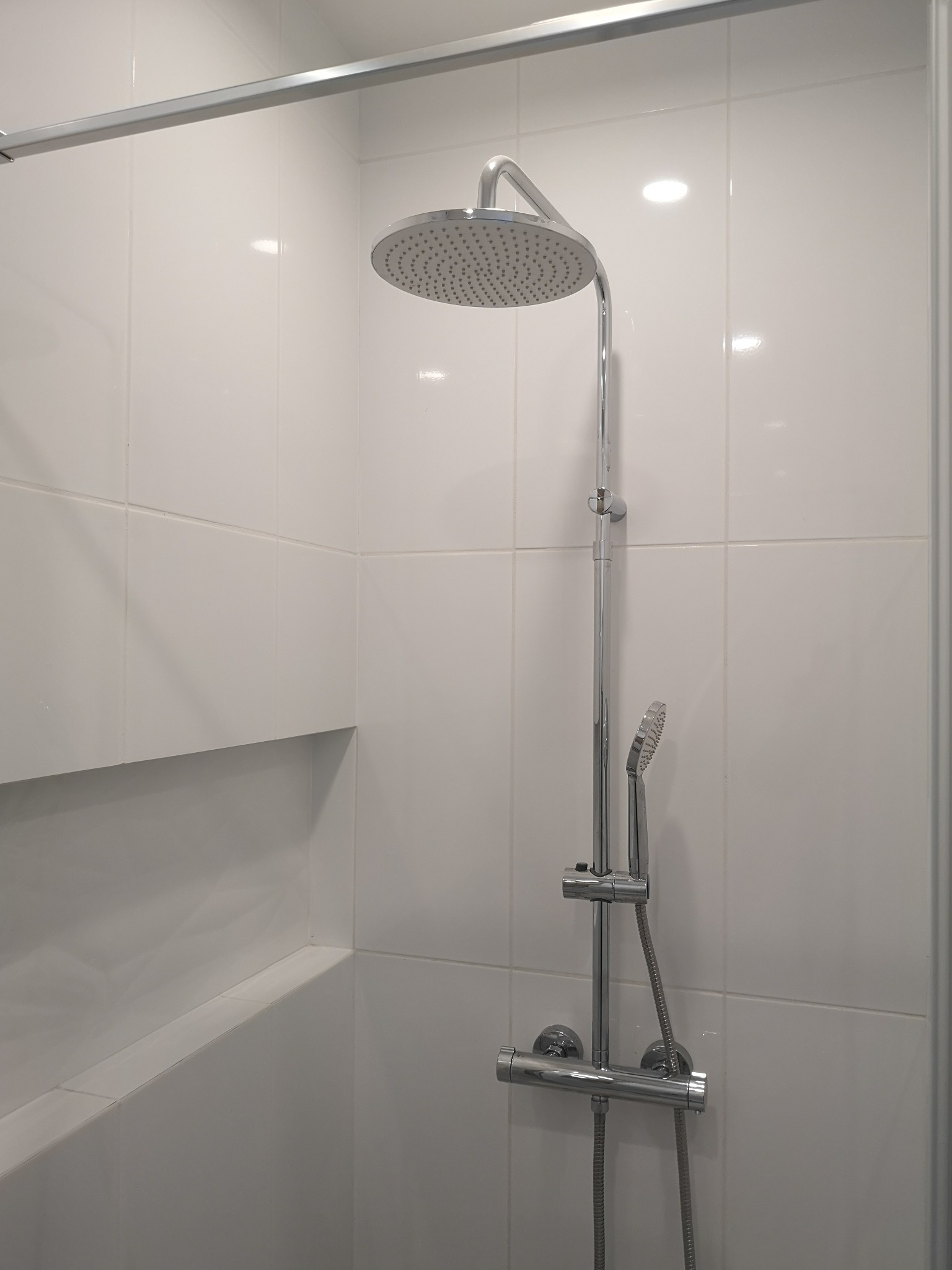 Nowoczesna kabina prysznicowa z chromowaną armaturą i deszczownicą, wykończona białymi, kwadratowymi płytkami ceramicznymi i wnęką na kosmetyki.