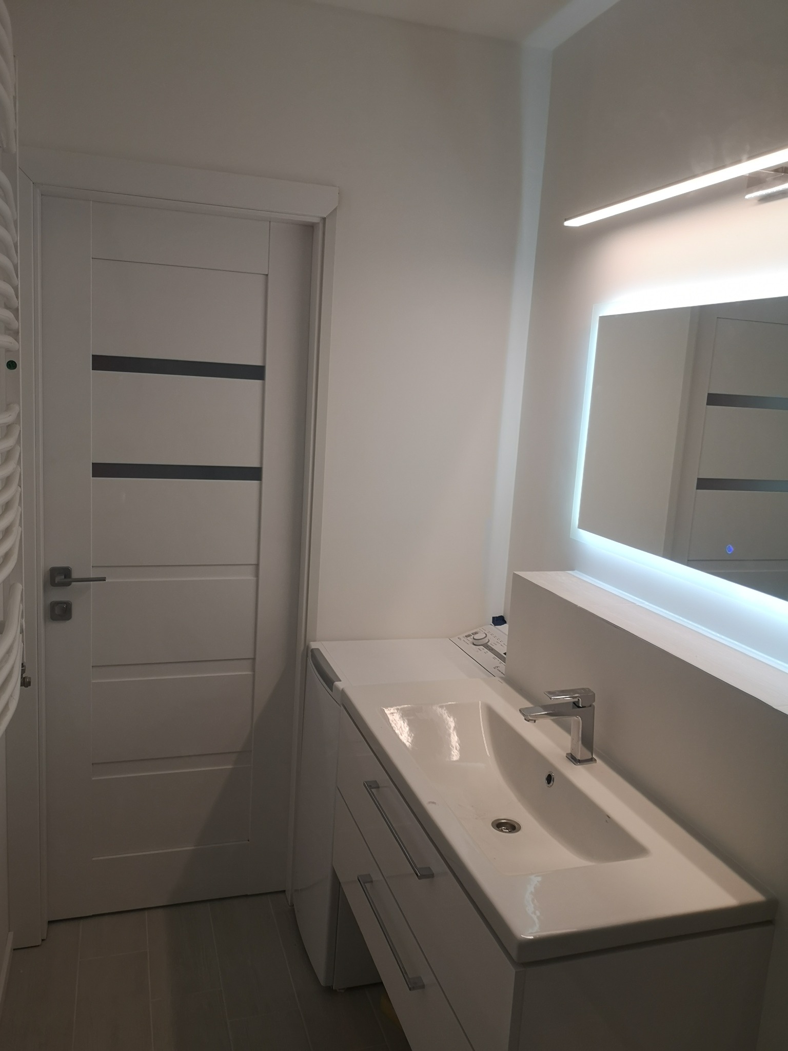 Nowoczesna, biała łazienka z umywalką nablatową, podświetlanym lustrem LED i drzwiami z poziomymi wstawkami w odcieniach szarości.