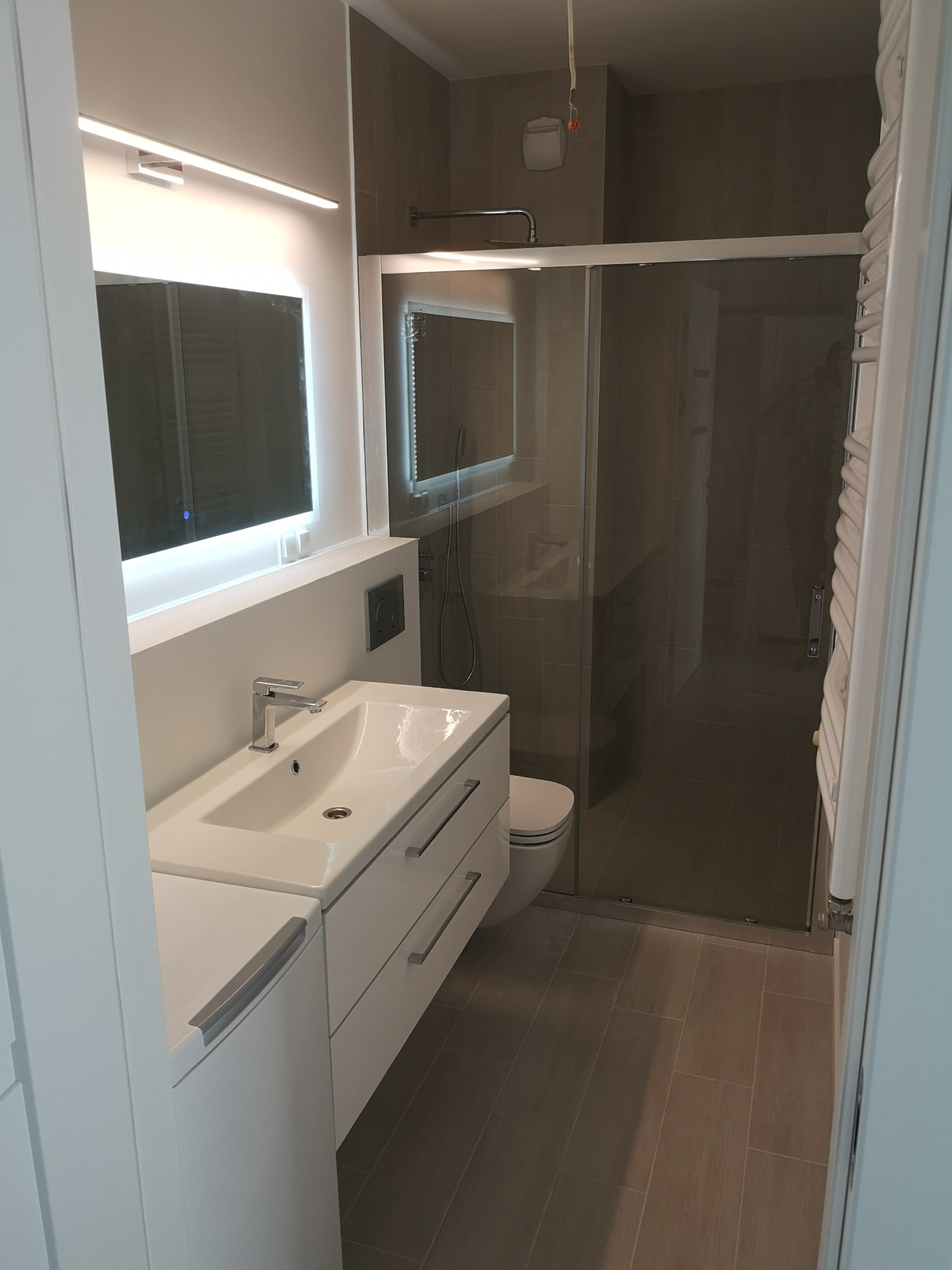 Nowoczesna łazienka z umywalką w zabudowie, podświetlanym lustrem i kabiną prysznicową z przesuwnymi drzwiami, wykończona płytkami w odcieniach szarości.