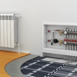 Biały grzejnik panelowy na ścianie obok rozdzielacza ogrzewania podłogowego z pętlami rur w szarym betonie, częściowo przykrytymi drewnianą podłogą.