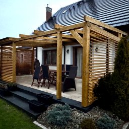 Tom Art Tomasz Kowaski - Drewniana pergola z poziomymi lamelami osłaniająca taras z meblami ogrodowymi, widok na dom z ciemną dachówką.