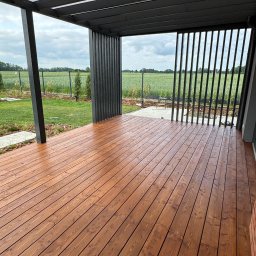 Tom Art Tomasz Kowaski - Nowoczesny taras z drewna w ciepłym odcieniu brązu, z czarną pergolą i widokiem na zielony krajobraz. Minimalistyczny design i naturalne materiały.