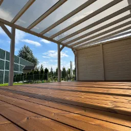 Drewniany taras z zadaszeniem z przezroczystych paneli, widok z dołu, ciepłe oświetlenie, elementy ogrodzenia w tle, regularny układ desek.