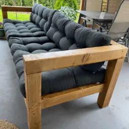 Trzyosobowa sofa z jasnego drewna z szarymi poduszkami na siedzisku i oparciu, ustawiona na szarym tarasie z widokiem na zielony ogród i zadaszoną część z meblami ogrodowymi.