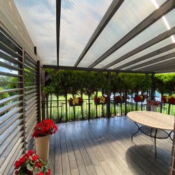 Tom Art Tomasz Kowaski - Zadaszona przestrzeń wypoczynkowa z drewnianą konstrukcją, przezroczystym zadaszeniem, kompozytową podłogą, kwiatami w donicach i okrągłym stołem z mozaikowym blatem.