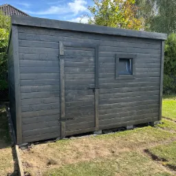 Ciemna, drewniana konstrukcja z oknem i drzwiami, prawdopodobnie domek narzędziowy, ustawiona na betonowych blokach na trawniku w słoneczny dzień.