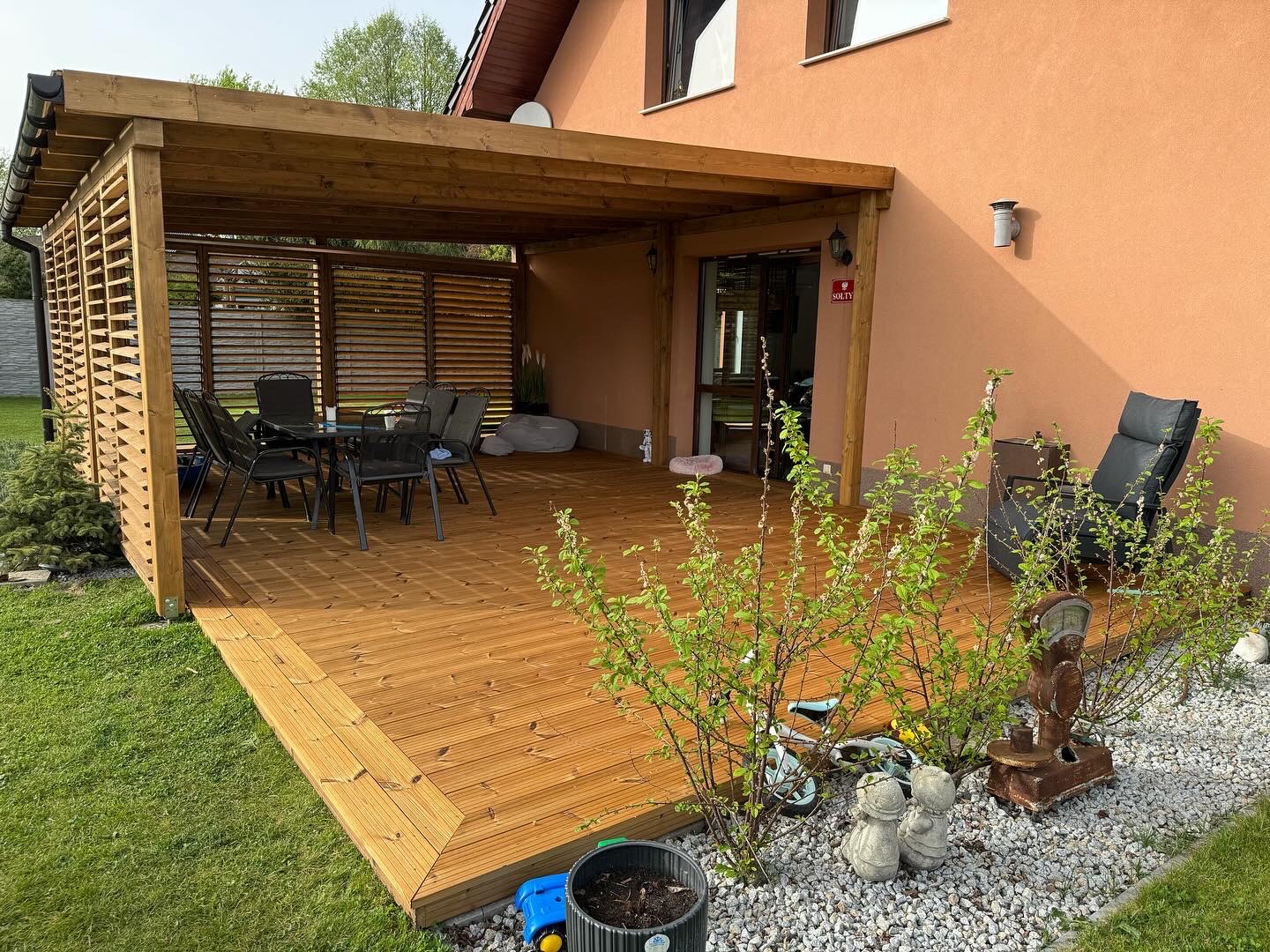 Zadaszony taras z drewna z ażurowymi ścianami, stół z krzesłami, leżak, dekoracje ogrodowe na żwirowym podłożu, w tle ściana budynku w kolorze terakoty.