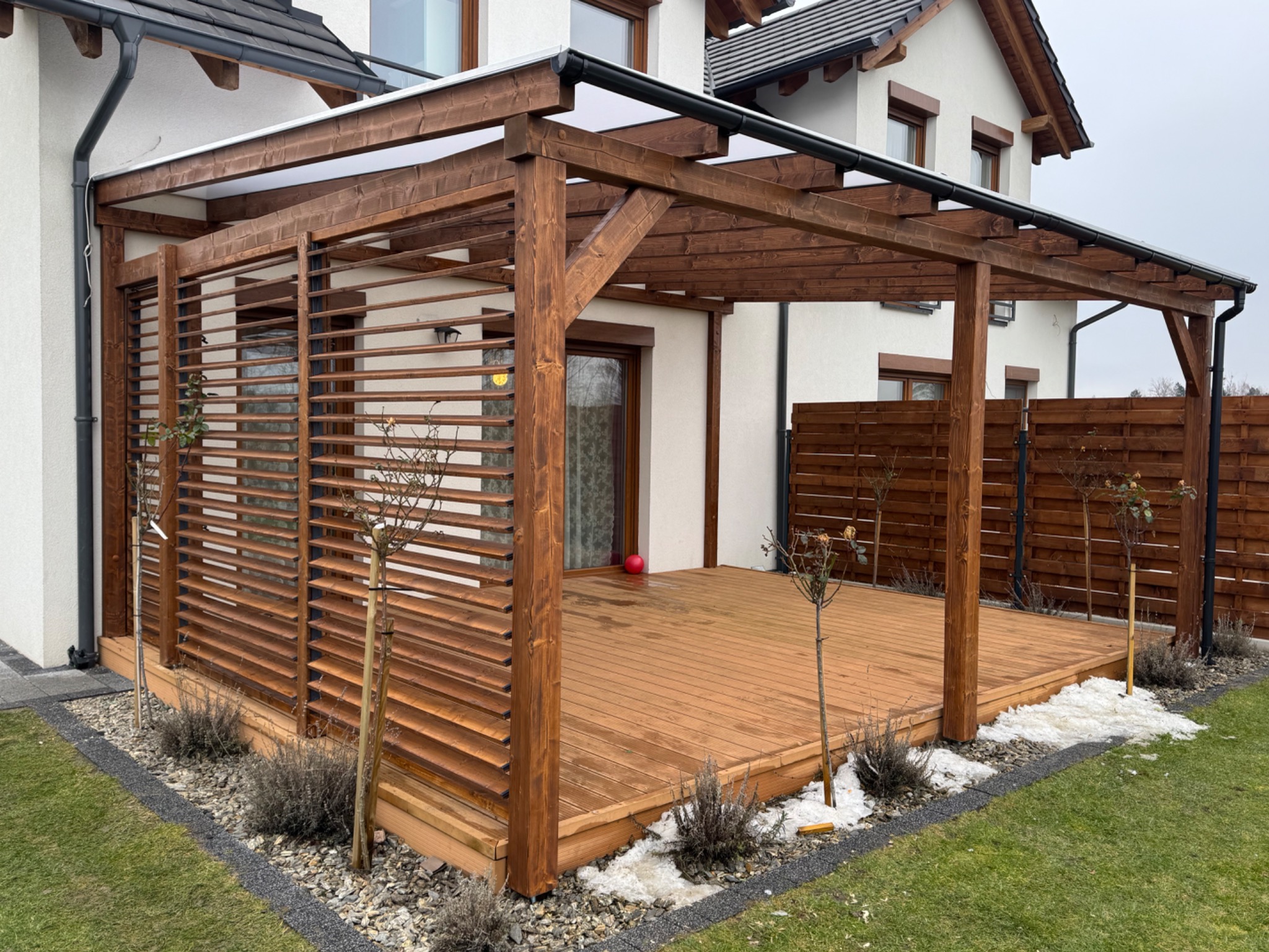 Zadaszona konstrukcja z drewna z przesłoniętą ścianą boczną, połączona z tarasem, z widocznymi elementami ogrodu w zimowej scenerii.