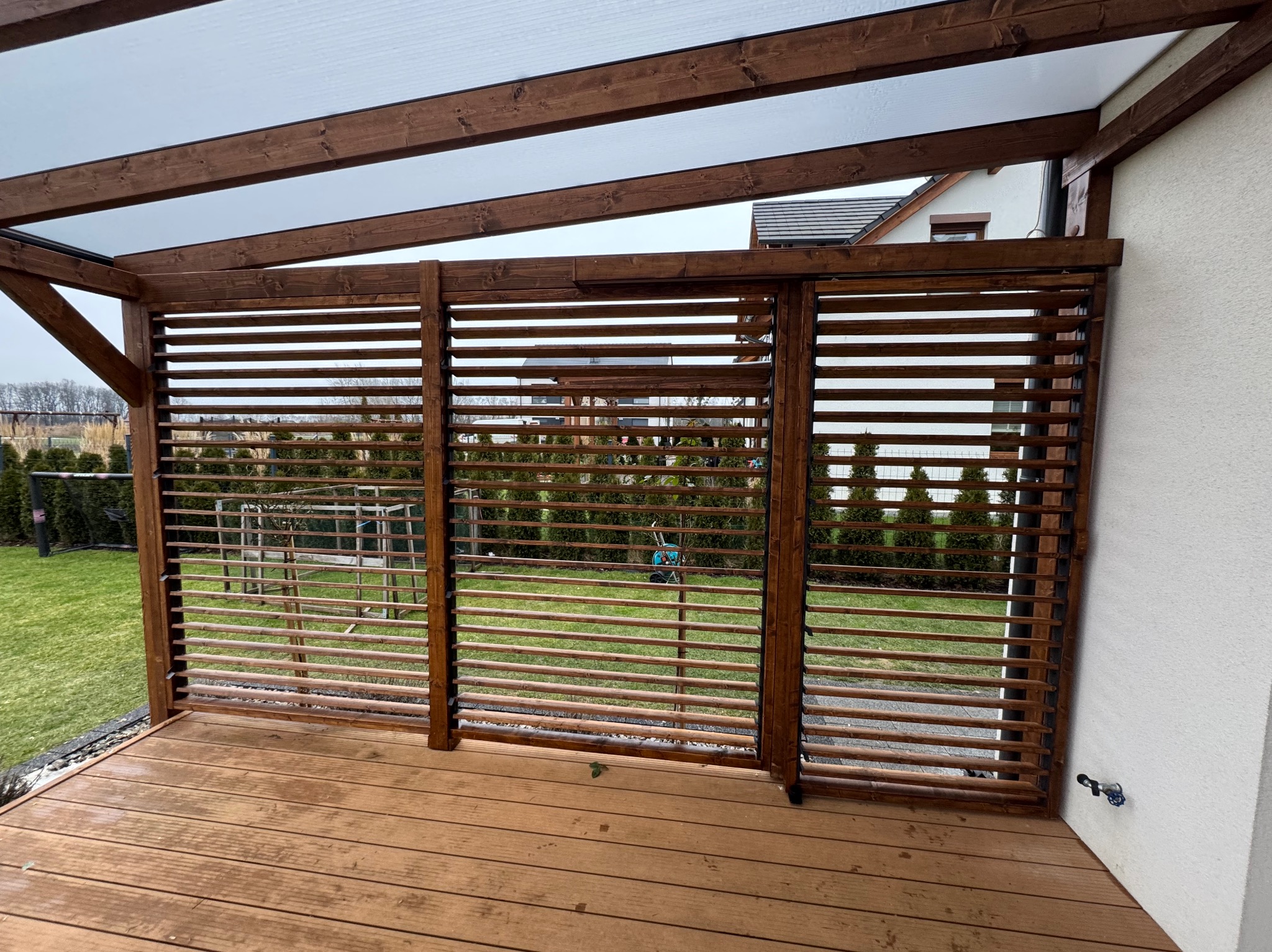 Zadaszona konstrukcja z drewnianymi lamelami tworzącymi ścianę, widok na ogród z zielonym trawnikiem, drewniana podłoga tarasu, biała ściana budynku z widoczną instalacją.