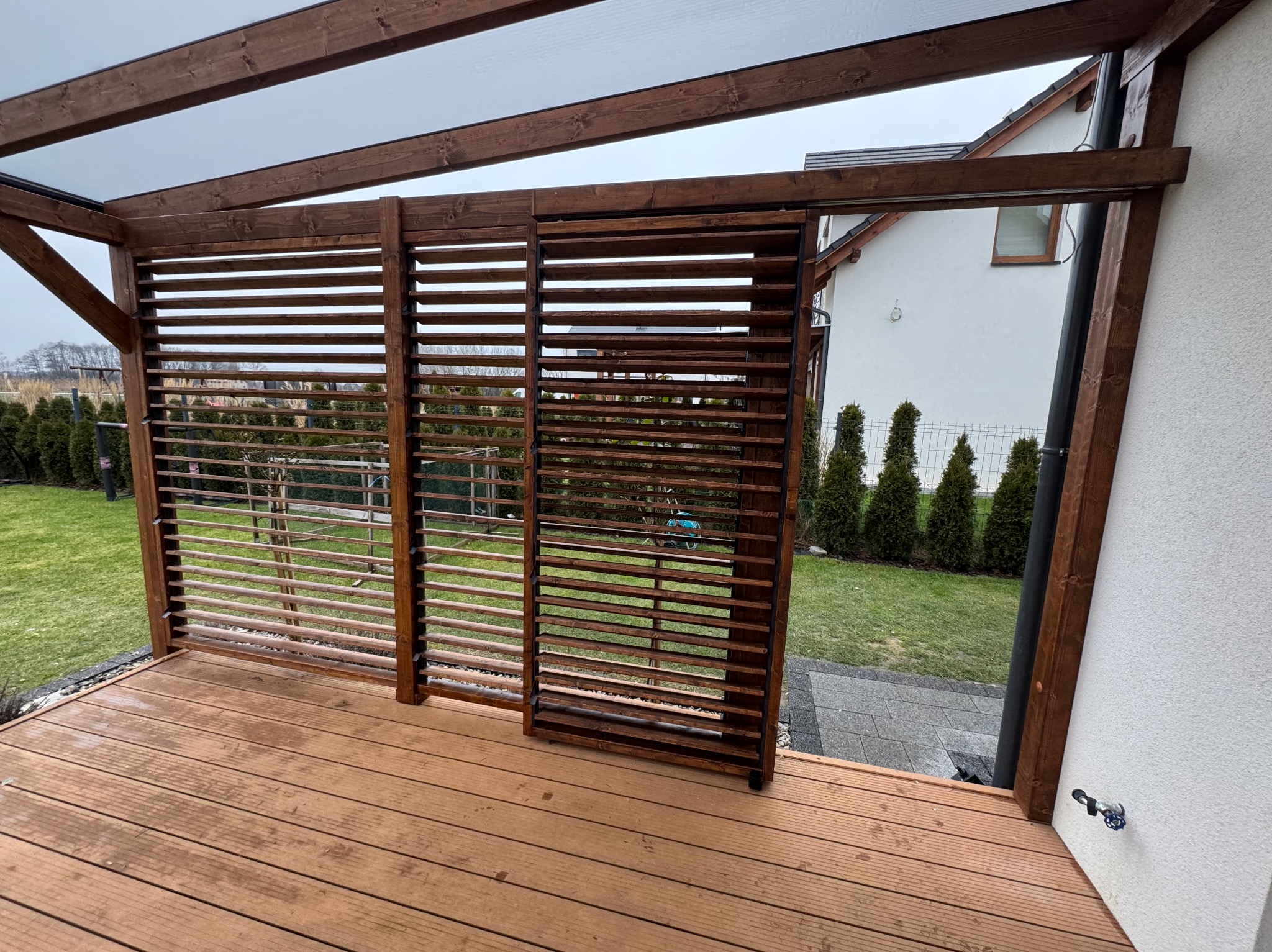 Zadaszona konstrukcja z drewnianymi żaluzjami i drewnianym tarasem, widok na trawnik i budynek mieszkalny w tle.