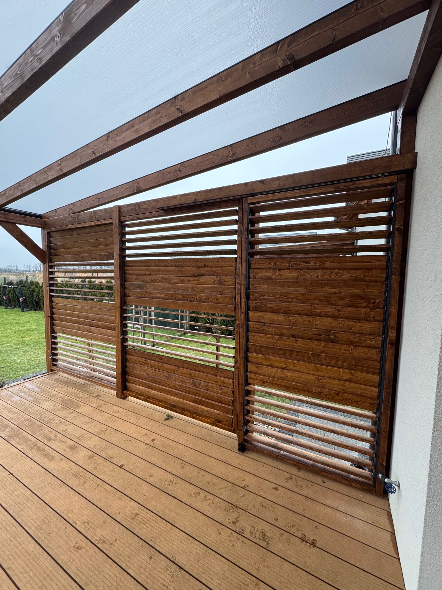 Zadaszony taras drewniany z drewnianymi, przesuwnymi panelami osłonowymi, widok na ogród przez szczeliny w panelach, drewniana podłoga z widocznymi śladami użytkowania.