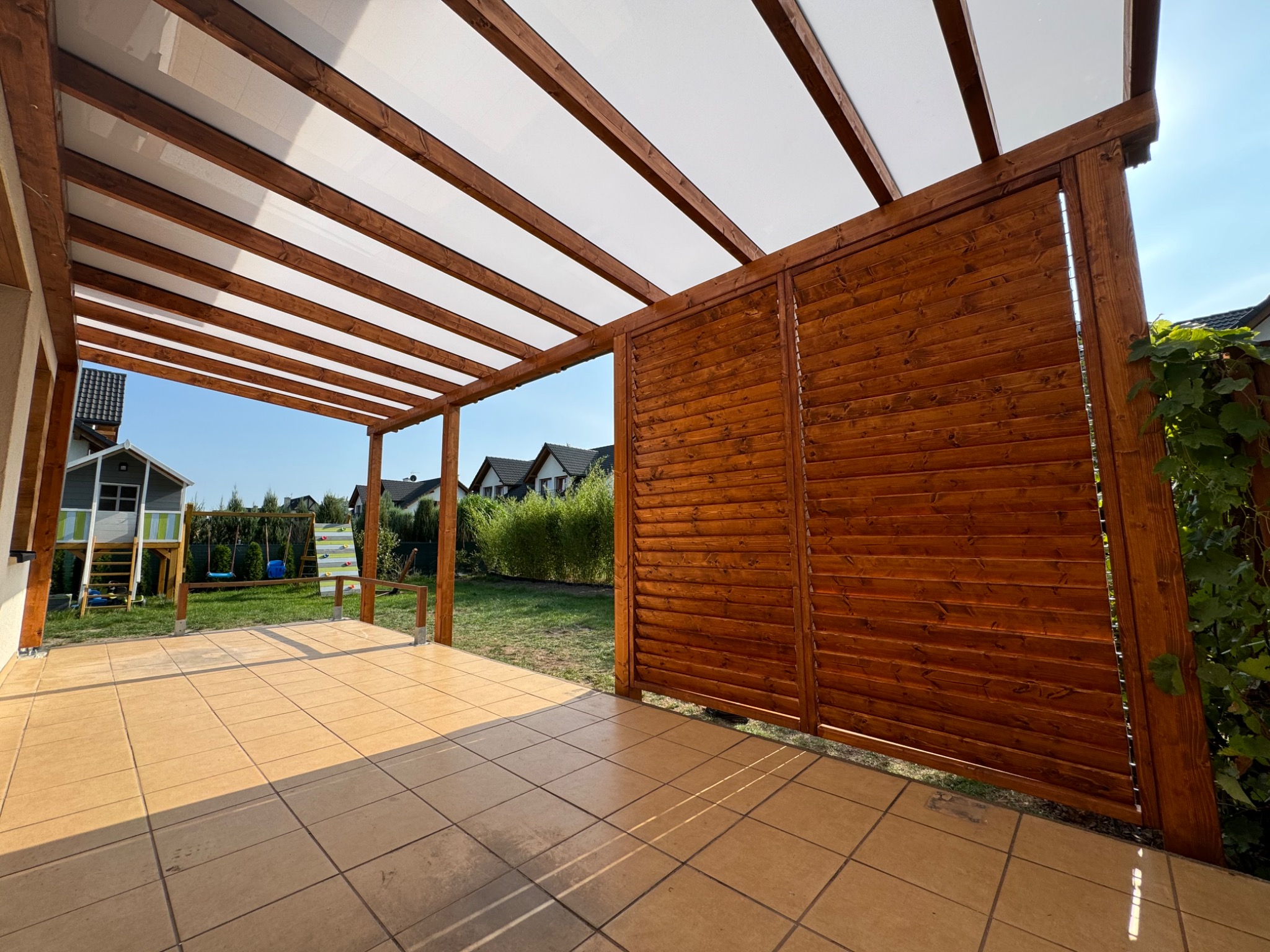 Zadaszona konstrukcja z drewna z ażurową ścianką boczną, osłaniająca część tarasu z jasnobrązowych płytek. W tle widoczny plac zabaw i zabudowa jednorodzinna.