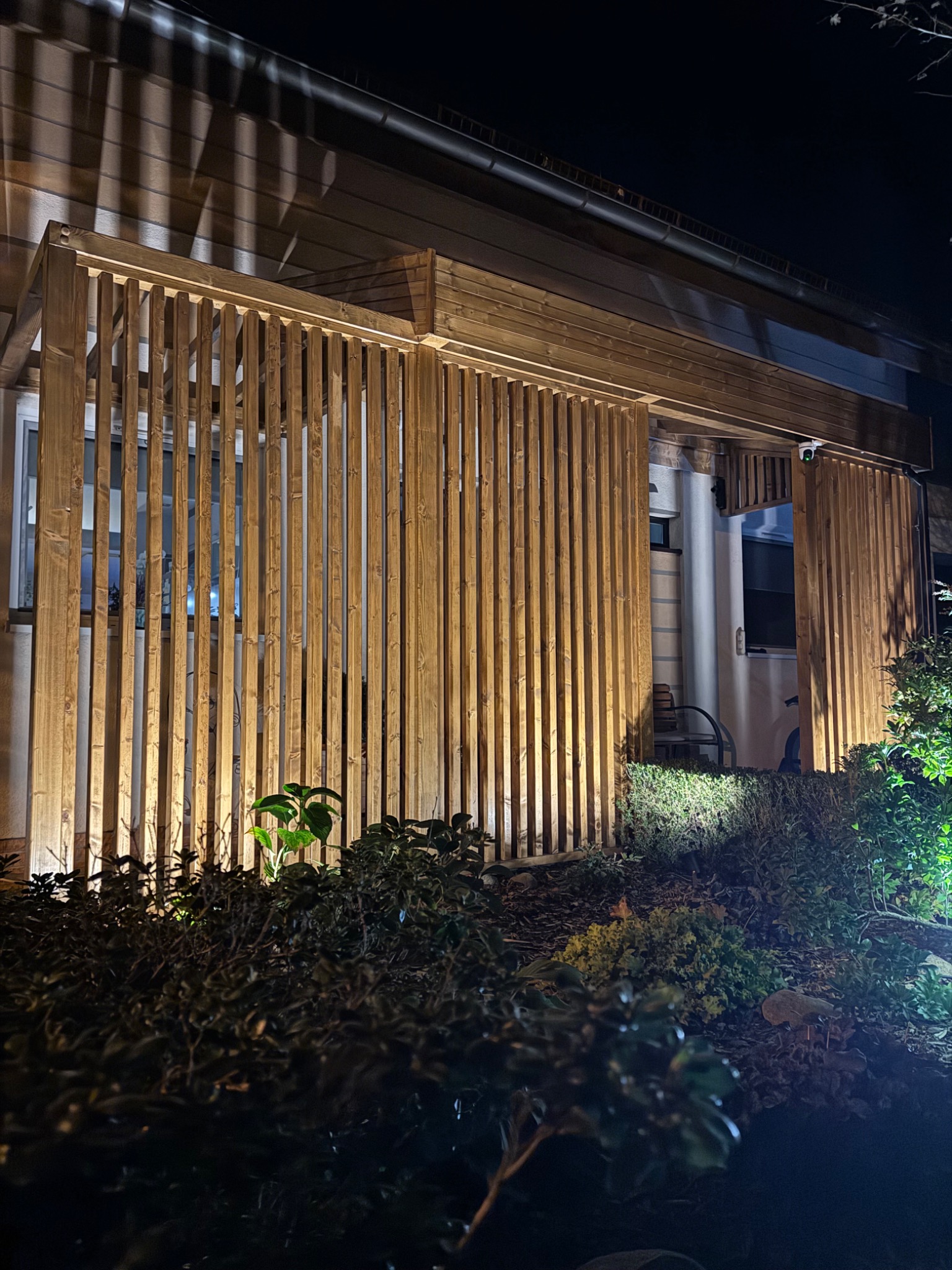 Drewniana konstrukcja z pionowych listew, oświetlona od góry i dołu, tworząca przesłonę przed domem, widoczna nocą, z roślinnością w tle.