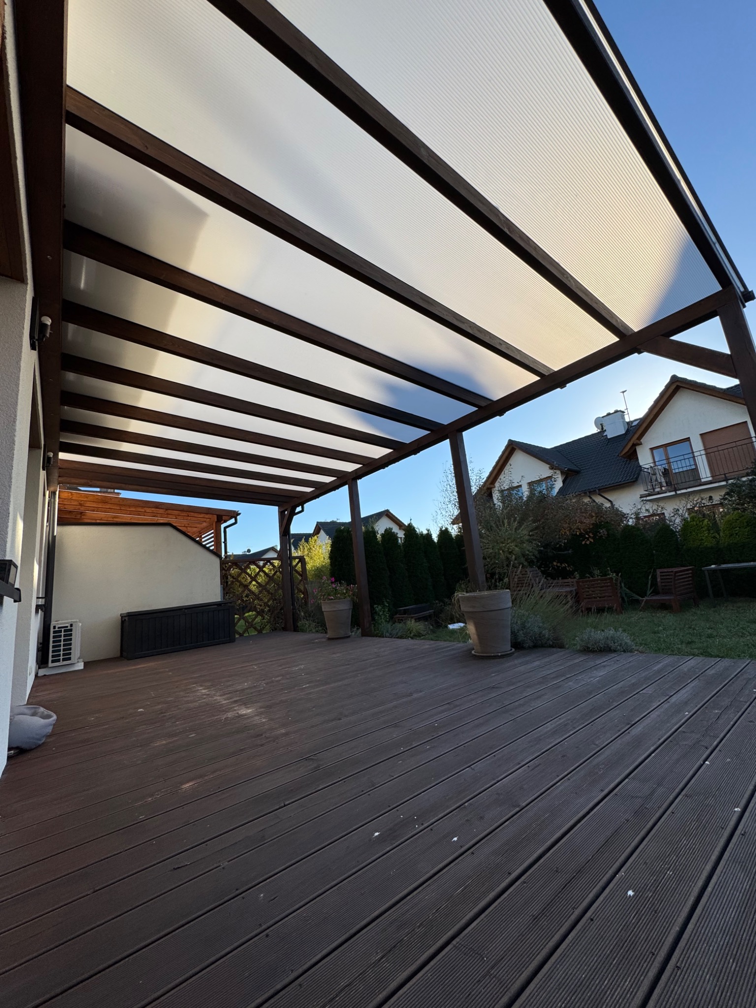 Zadaszony taras z brązowych desek kompozytowych, widok z dołu na białe zadaszenie i drewniane belki, w tle dom z ogrodem.