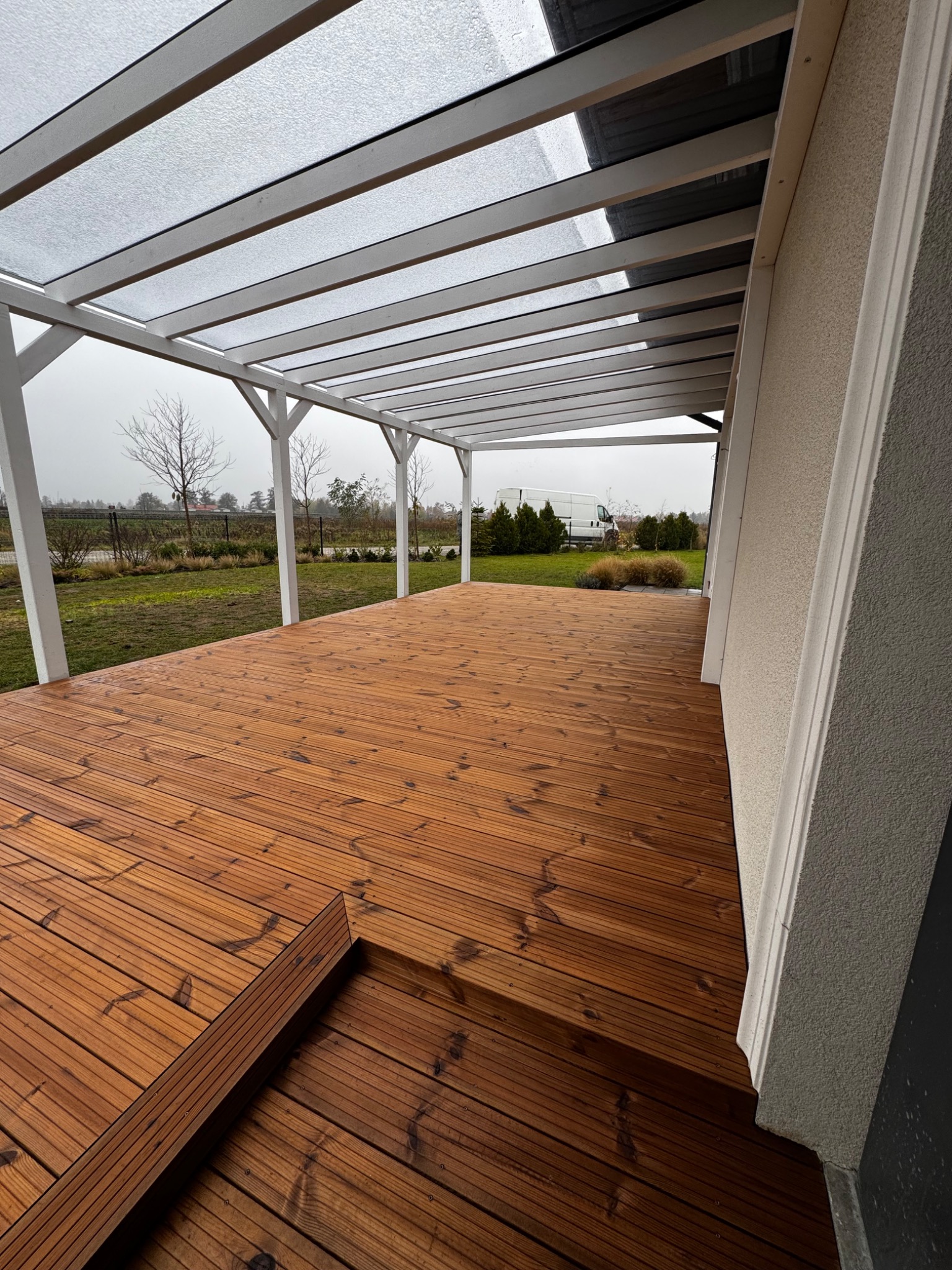 Nowoczesny taras z drewna zadaszony pergolą z białymi belkami, widok na ogród w pochmurny dzień, z elementami schodów.