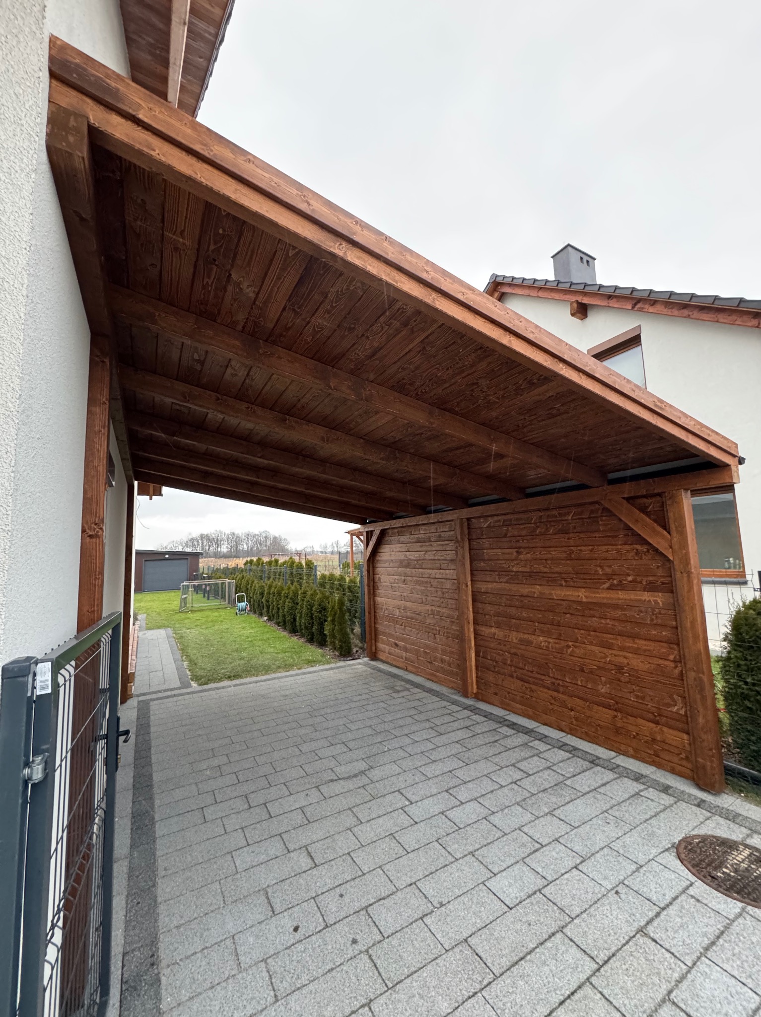 Zadaszone miejsce parkingowe z drewnianą konstrukcją i ścianą boczną, wykończone ciemnym brązem, na tle domu z szarym dachem i starannie przystrzyżonym żywopłotem.