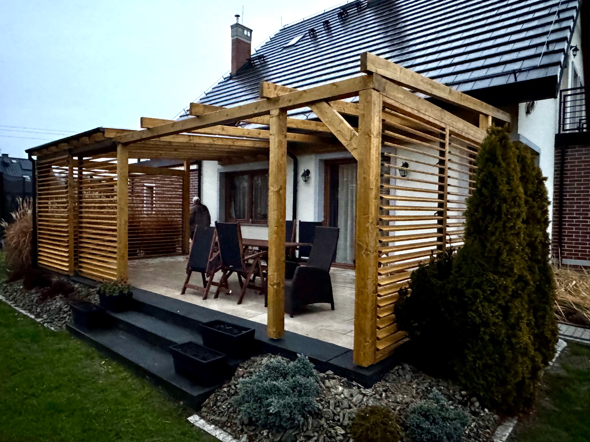 Drewniana pergola z poziomymi lamelami osłaniająca taras z meblami ogrodowymi, widok na dom z ciemną dachówką.