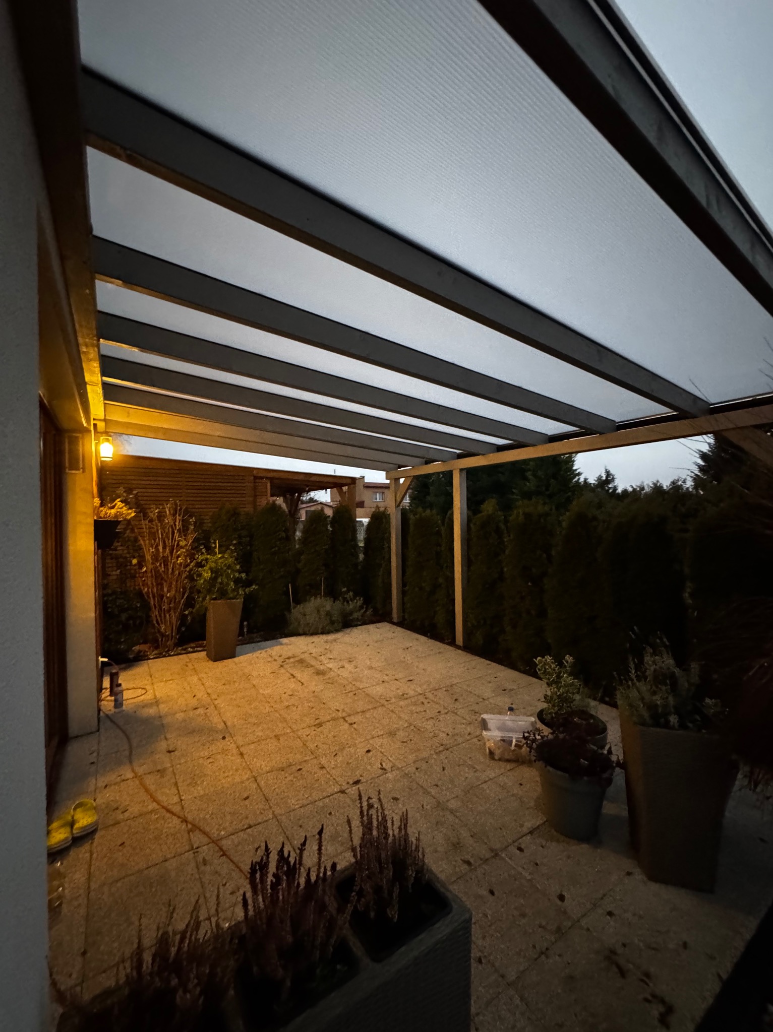 Zadaszenie tarasu z poliwęglanu i ciemnej konstrukcji ramowej, widok na ogród z tujami i drewnianym płotem w tle, oświetlone ciepłym światłem z budynku.