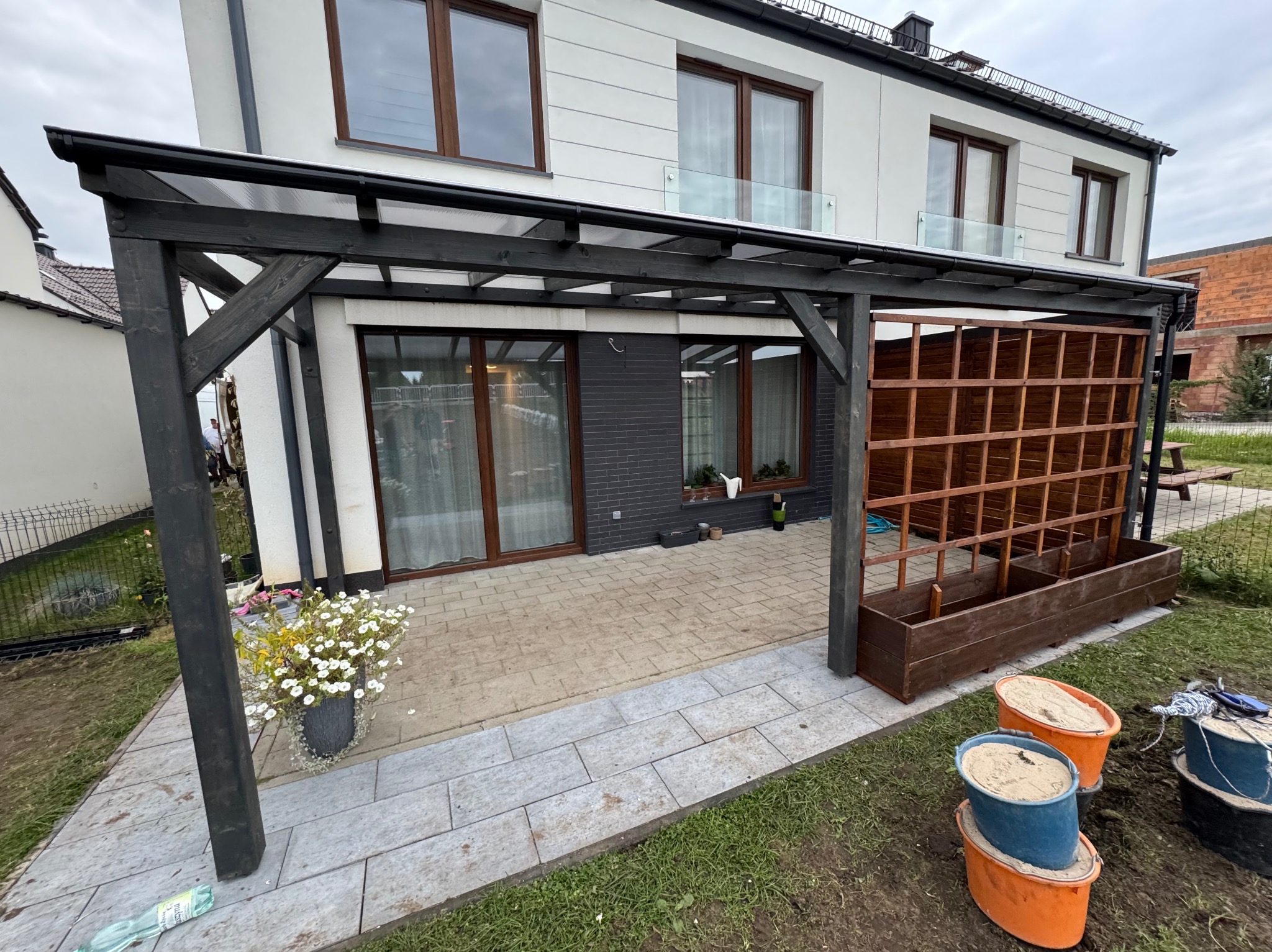 Ciemna, drewniana konstrukcja z zadaszeniem przylegająca do domu, z drewnianą ścianką ażurową, donicą z kwiatami i wiadrami z piaskiem obok, na wyłożonym płytami tarasie.