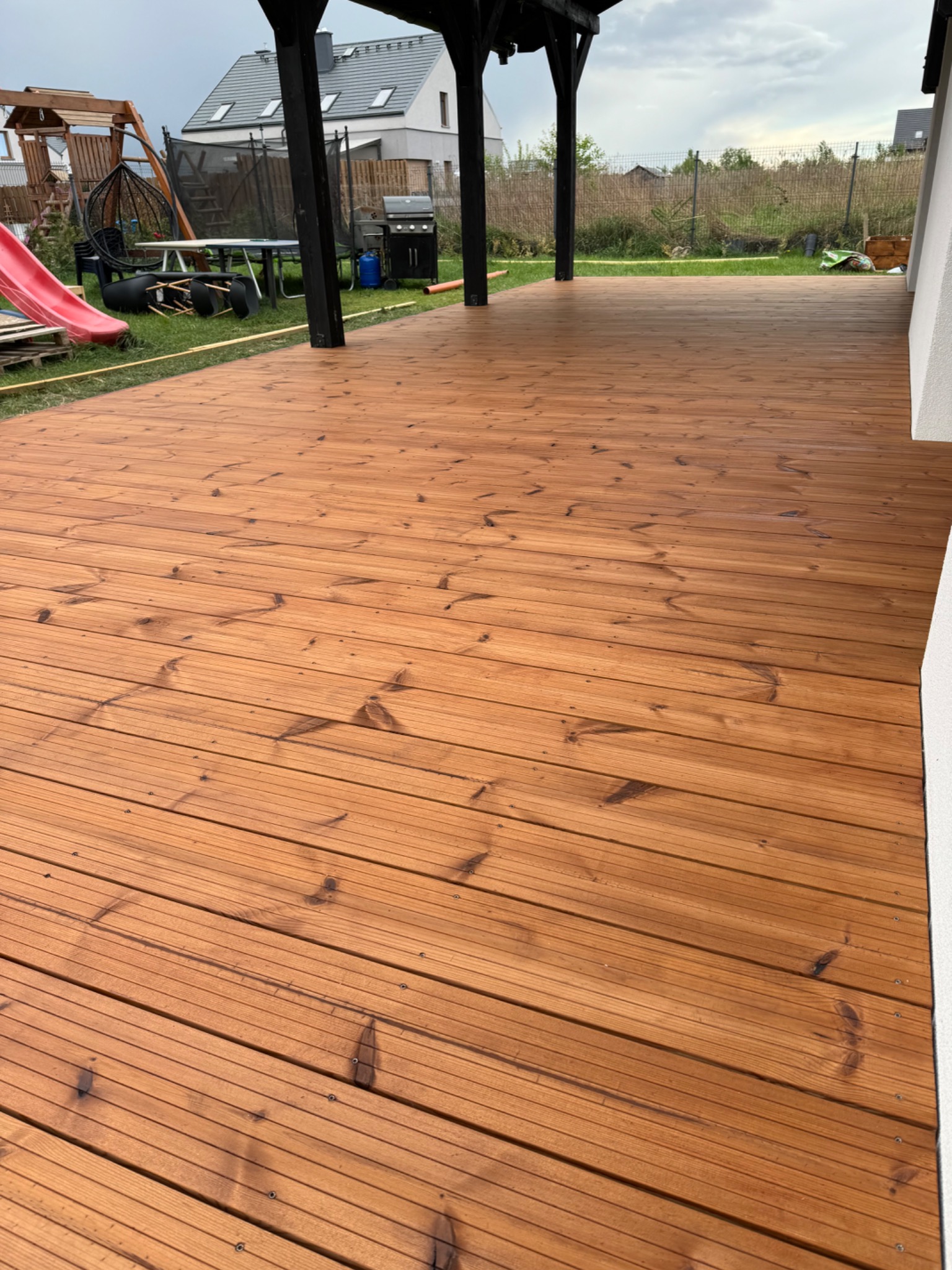 Nowo wybudowany taras z desek w kolorze miodowym, z czarną drewnianą konstrukcją zadaszenia, widok na trawnik z elementami placu zabaw i grilla, w tle nowoczesny dom.