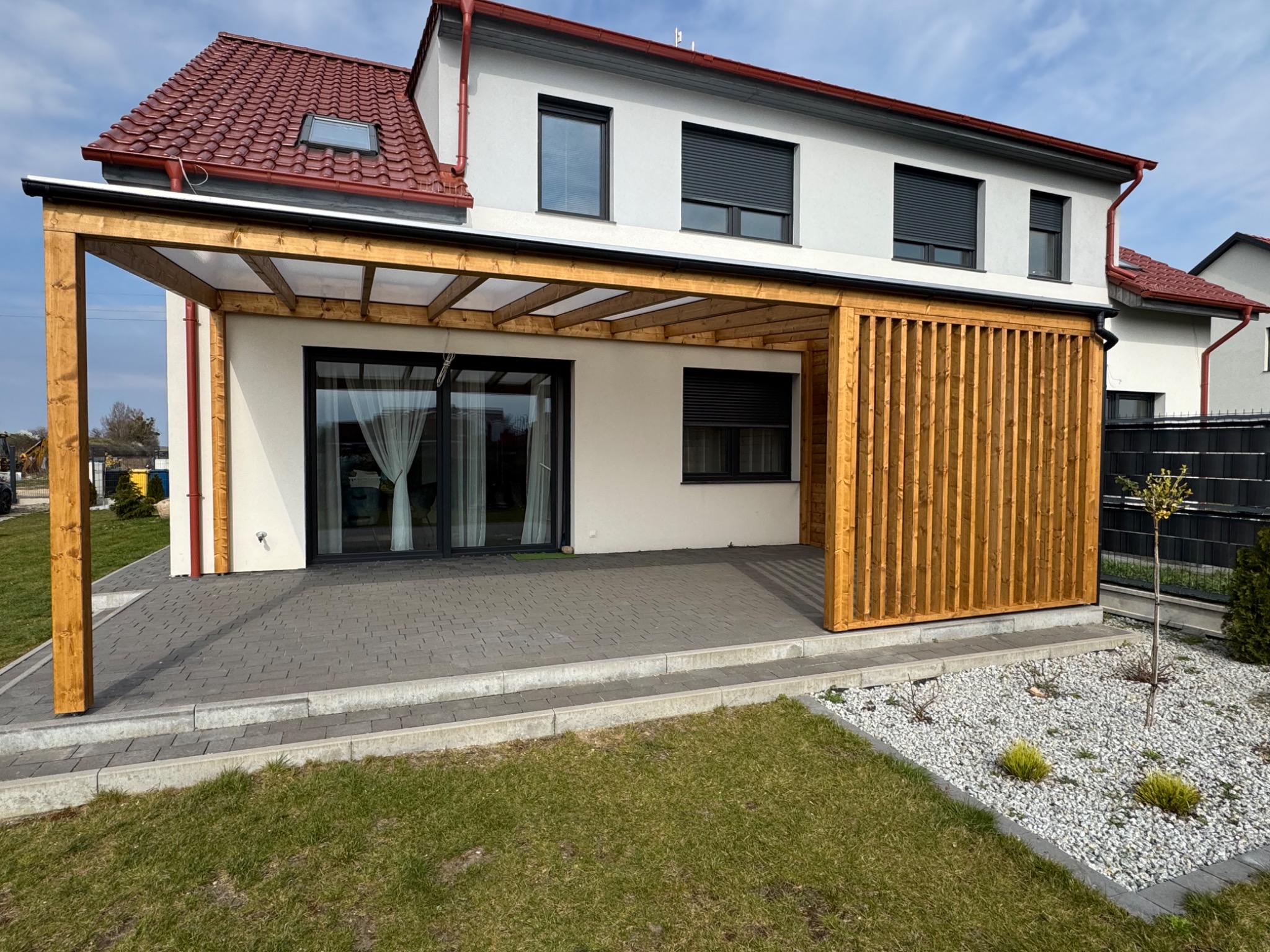 Nowoczesna konstrukcja z drewna, częściowo zadaszona, przylegająca do domu z dużymi oknami tarasowymi, z drewnianą ścianką działową z pionowych desek, wyłożona kostką brukową.