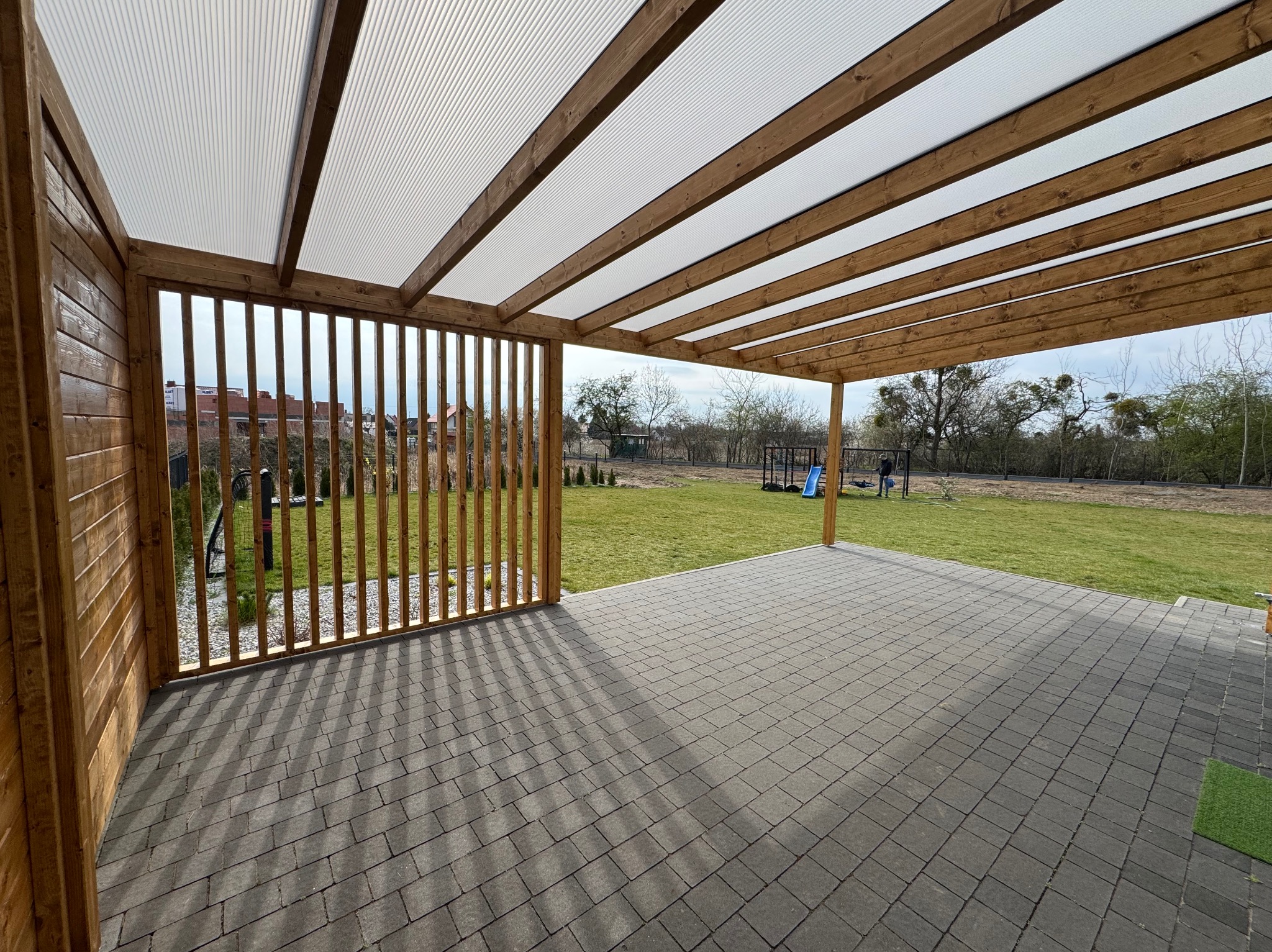 Zadaszona konstrukcja z drewna z przezroczystym dachem, widok na duży trawnik z placem zabaw w tle, podłoga wyłożona szarą kostką brukową.