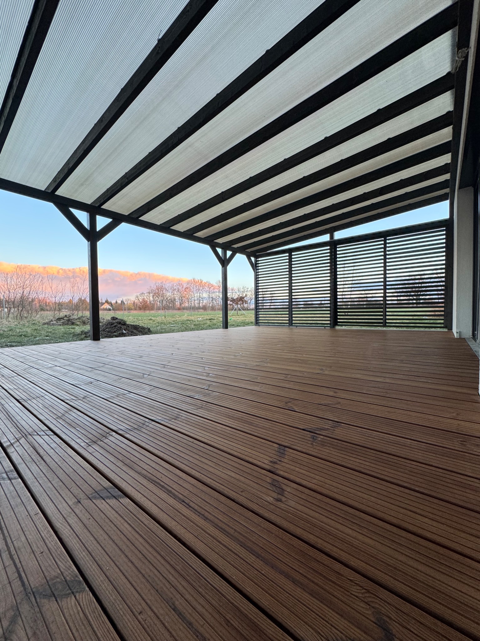 Drewniany taras z zadaszoną pergolą z przezroczystym dachem i drewnianymi żaluzjami bocznymi, widok na trawnik i drzewa w oddali.