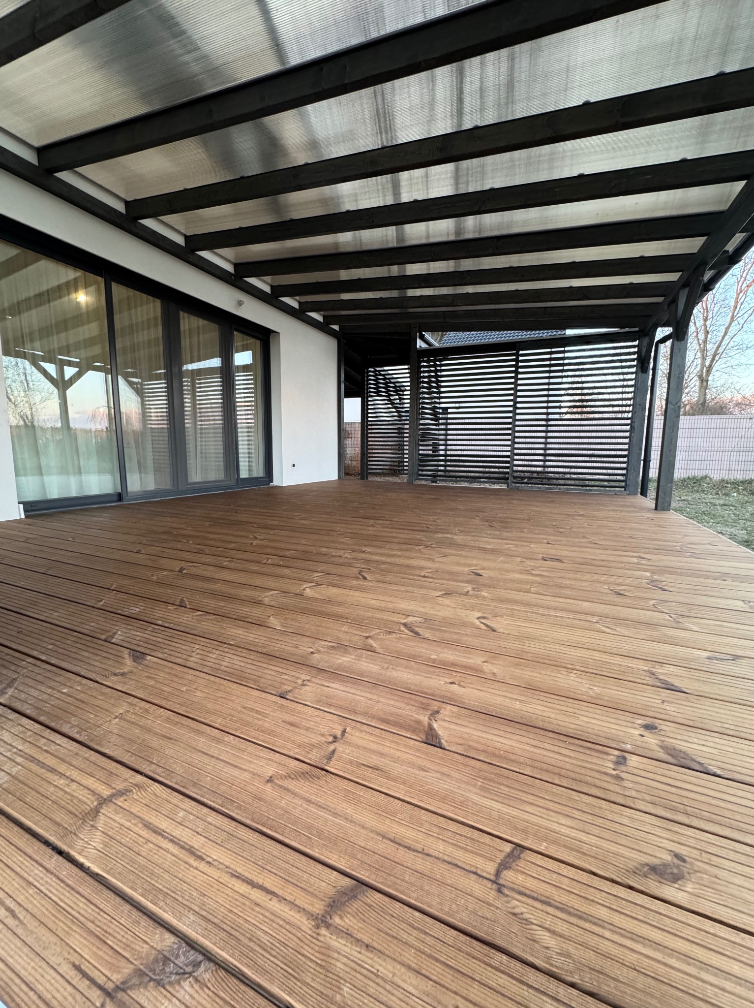 Przestronny taras z desek zadaszony konstrukcją z czarnych belek i przezroczystego zadaszenia, przylegający do nowoczesnego domu z dużymi przesuwnymi drzwiami.