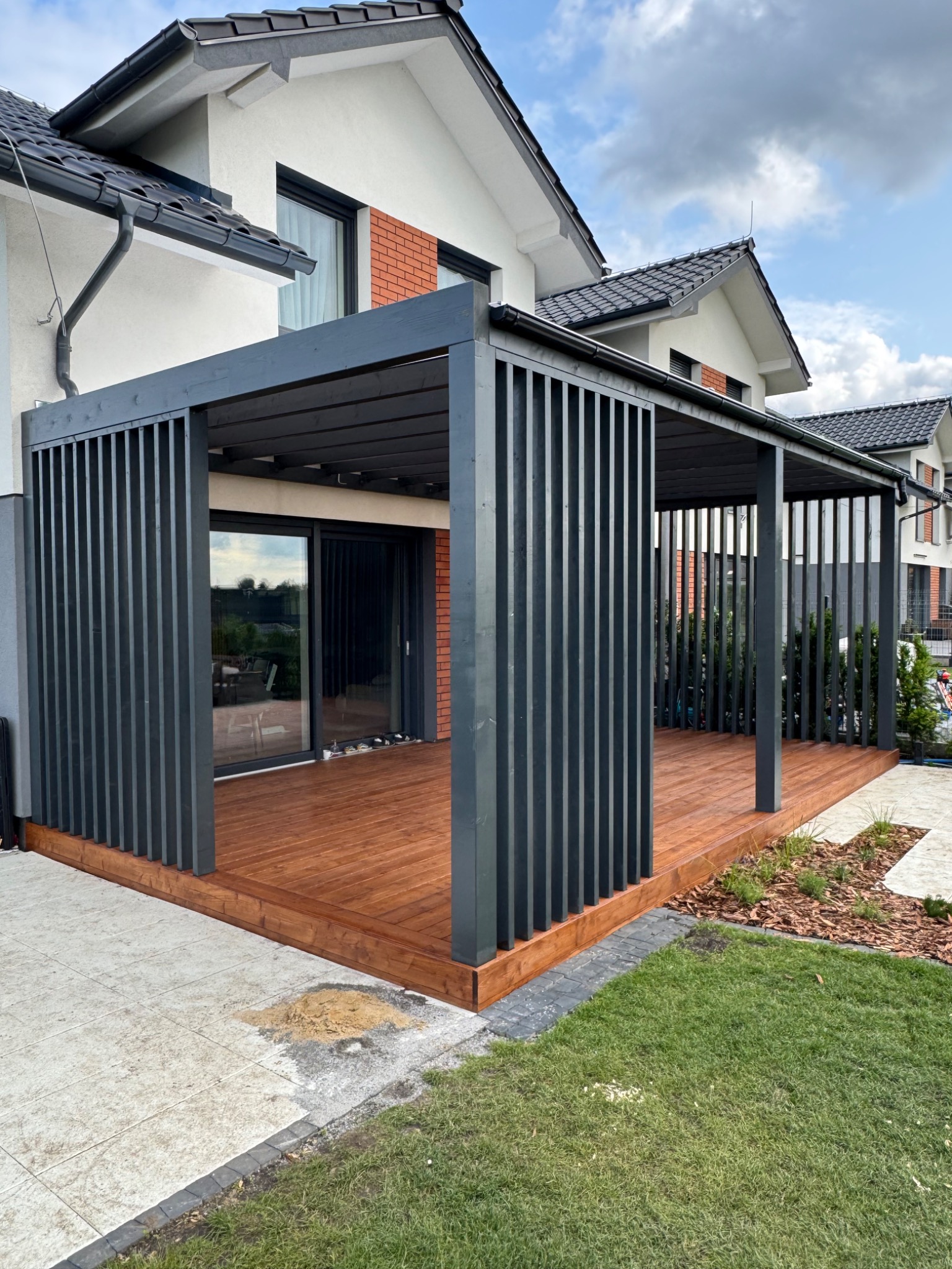 Nowoczesna pergola z ciemnoszarego drewna, połączona z tarasem z desek w ciepłym odcieniu brązu, przylegająca do jasnego domu z elementami cegły.