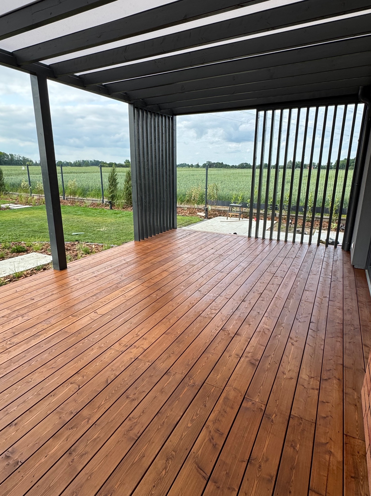 Nowoczesny taras z drewna w ciepłym odcieniu brązu, z czarną pergolą i widokiem na zielony krajobraz. Minimalistyczny design i naturalne materiały.