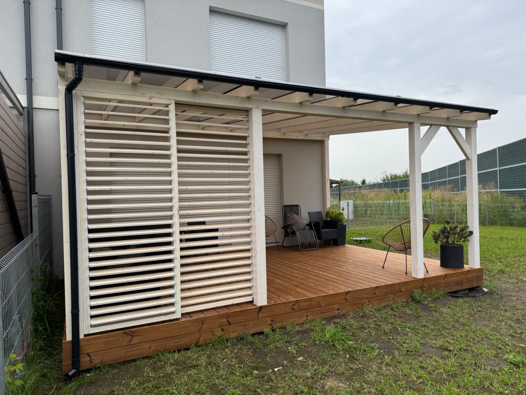 Drewniana konstrukcja z zadaszeniem i ażurową ścianą boczną, tworząca zadaszone miejsce wypoczynku na brązowym tarasie. Widoczne meble ogrodowe i rośliny w donicach.