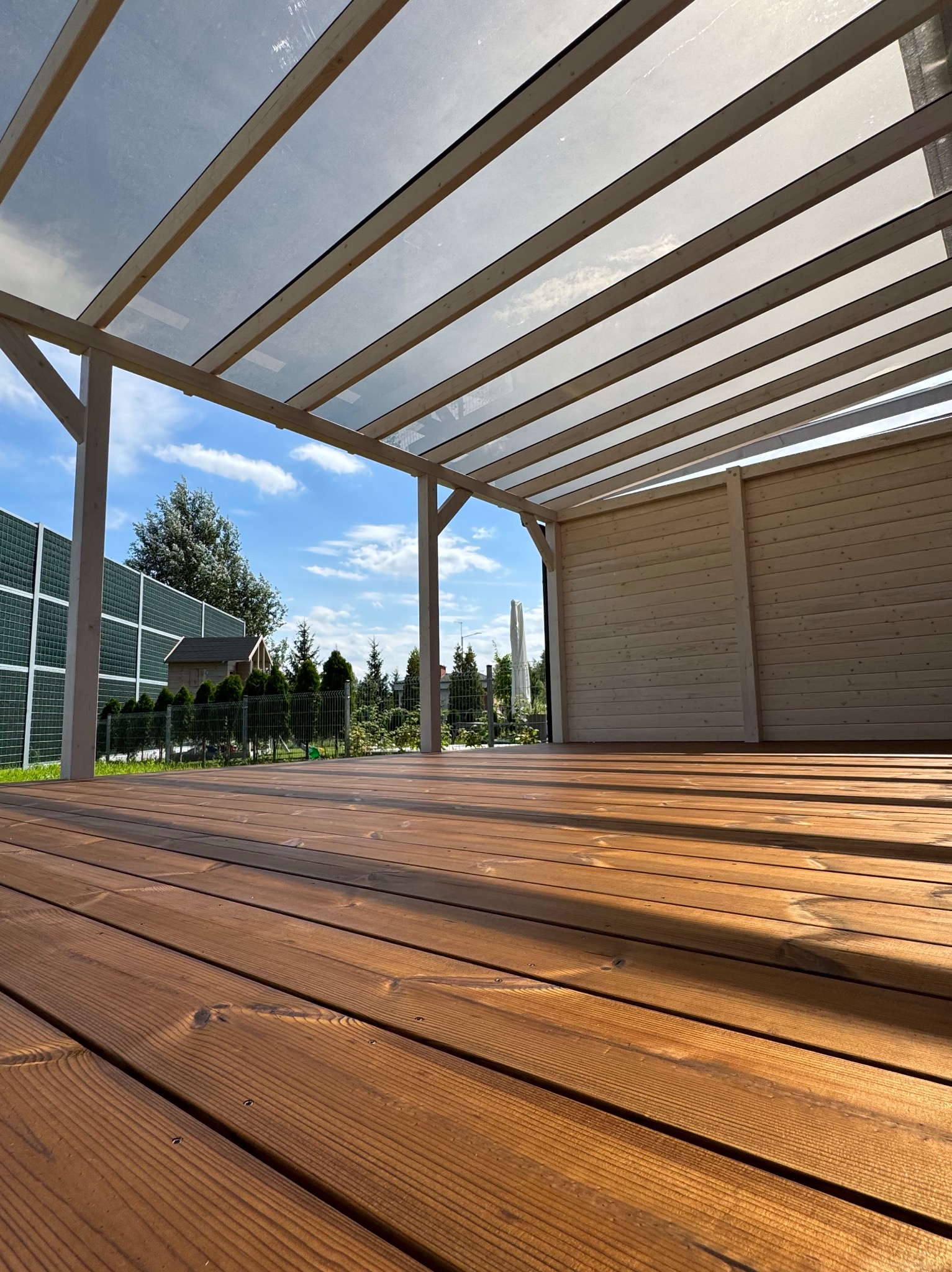 Drewniany taras z zadaszeniem z przezroczystych paneli, widok z dołu, ciepłe oświetlenie, elementy ogrodzenia w tle, regularny układ desek.