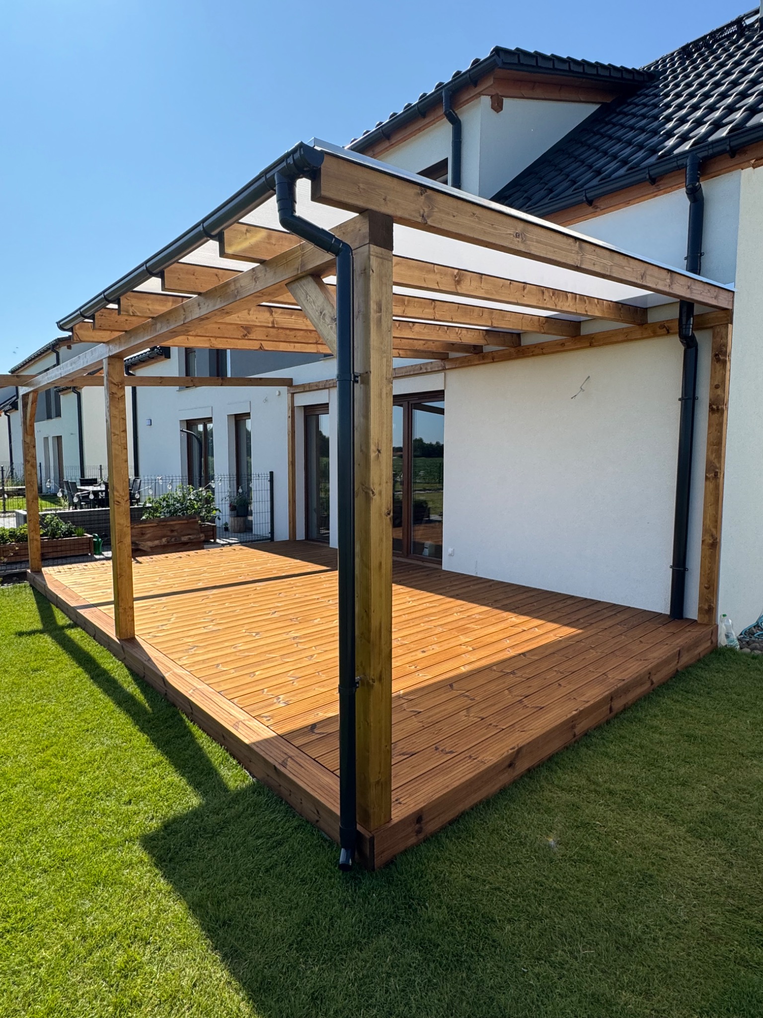 Drewniany taras z pergolą przy białym domu w słoneczny dzień. Widoczne deski tarasowe, drewniana konstrukcja pergoli i system odprowadzania wody.