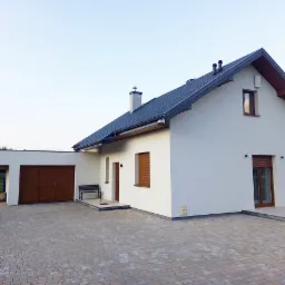 Nowoczesny dom jednorodzinny z białymi ścianami, ciemną dachówką i drewnianą bramą garażową, widok z podjazdu wyłożonego kostką brukową.