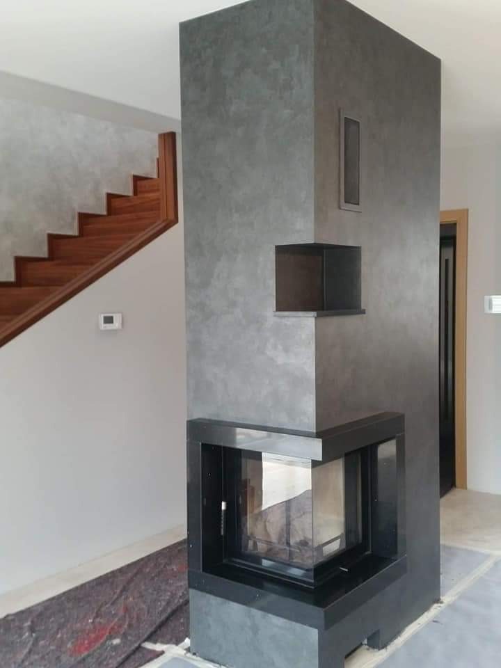 Nowoczesny kominek narożny z szarą, teksturowaną obudową, drewniane schody w tle, widoczne zabezpieczenie podłogi.