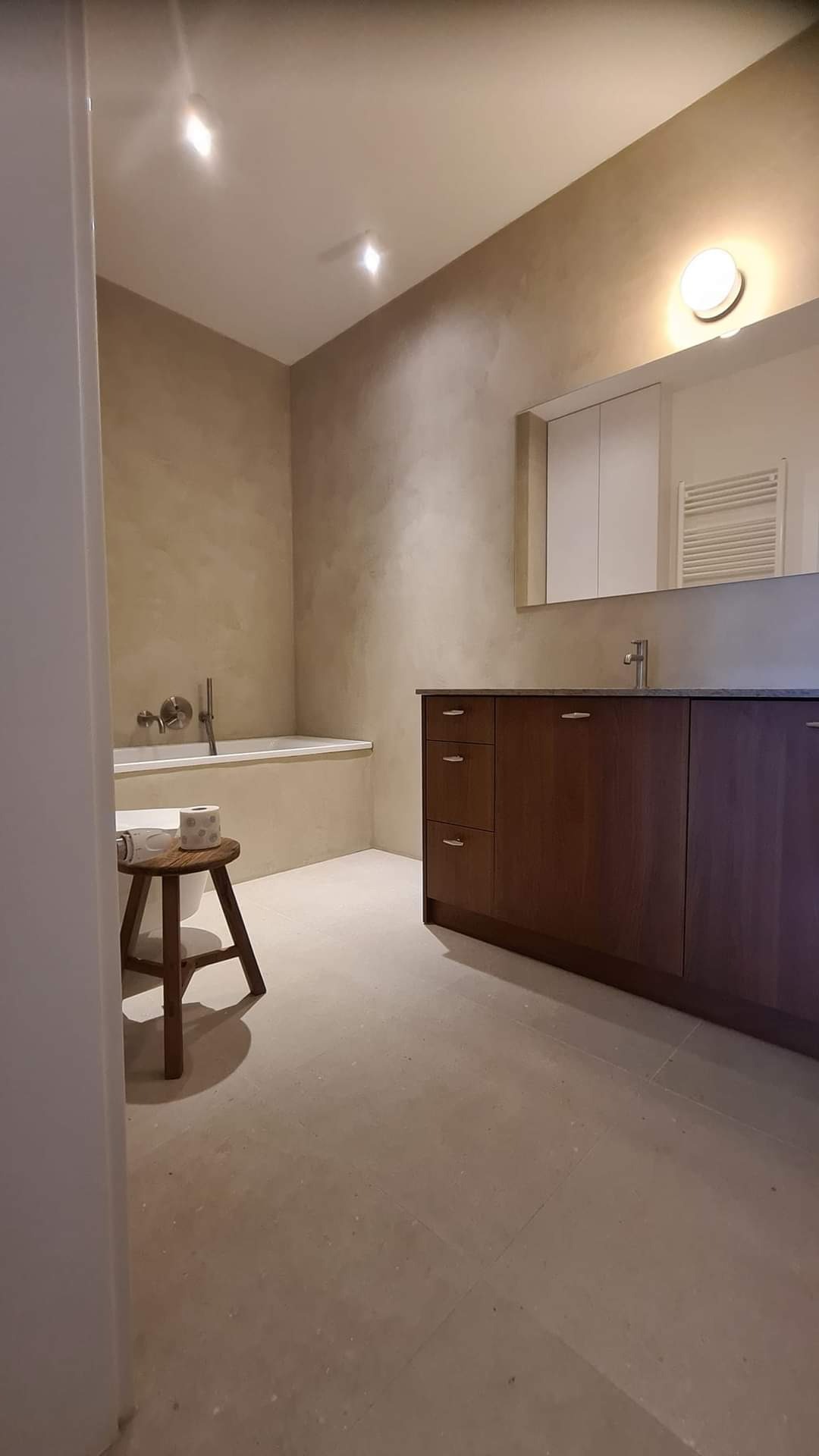 Wykończona łazienka z beżowymi ścianami i podłogą, drewniana szafka pod umywalką, wanna z baterią, drewniany stołek z papierem toaletowym.