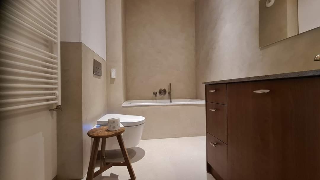 Łazienka wykończona mikrocementem w odcieniach beżu, z drewnianą szafką pod umywalką, grzejnikiem drabinkowym i toaletą z bidetem na tle wanny.