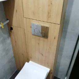 Toaleta w zabudowie z drewnopodobnej płyty z chromowanym przyciskiem spłukiwania na tle szarej ściany i podłogi.