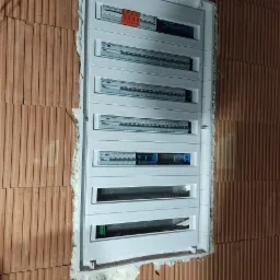 Rozdzielnica elektryczna natynkowa, zamontowana w surowej ścianie z cegły ceramicznej, zabezpieczona pianką montażową. Widoczne kable w górnej części ściany i niebieska rura karbowana w dolnej części.