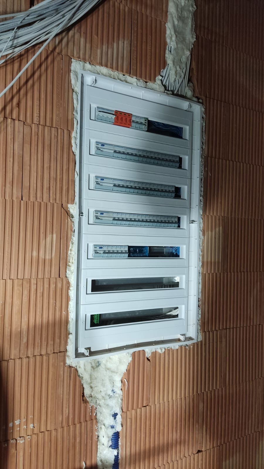 Rozdzielnica elektryczna natynkowa, zamontowana w surowej ścianie z cegły ceramicznej, zabezpieczona pianką montażową. Widoczne kable w górnej części ściany i niebieska rura karbowana w dolnej części.