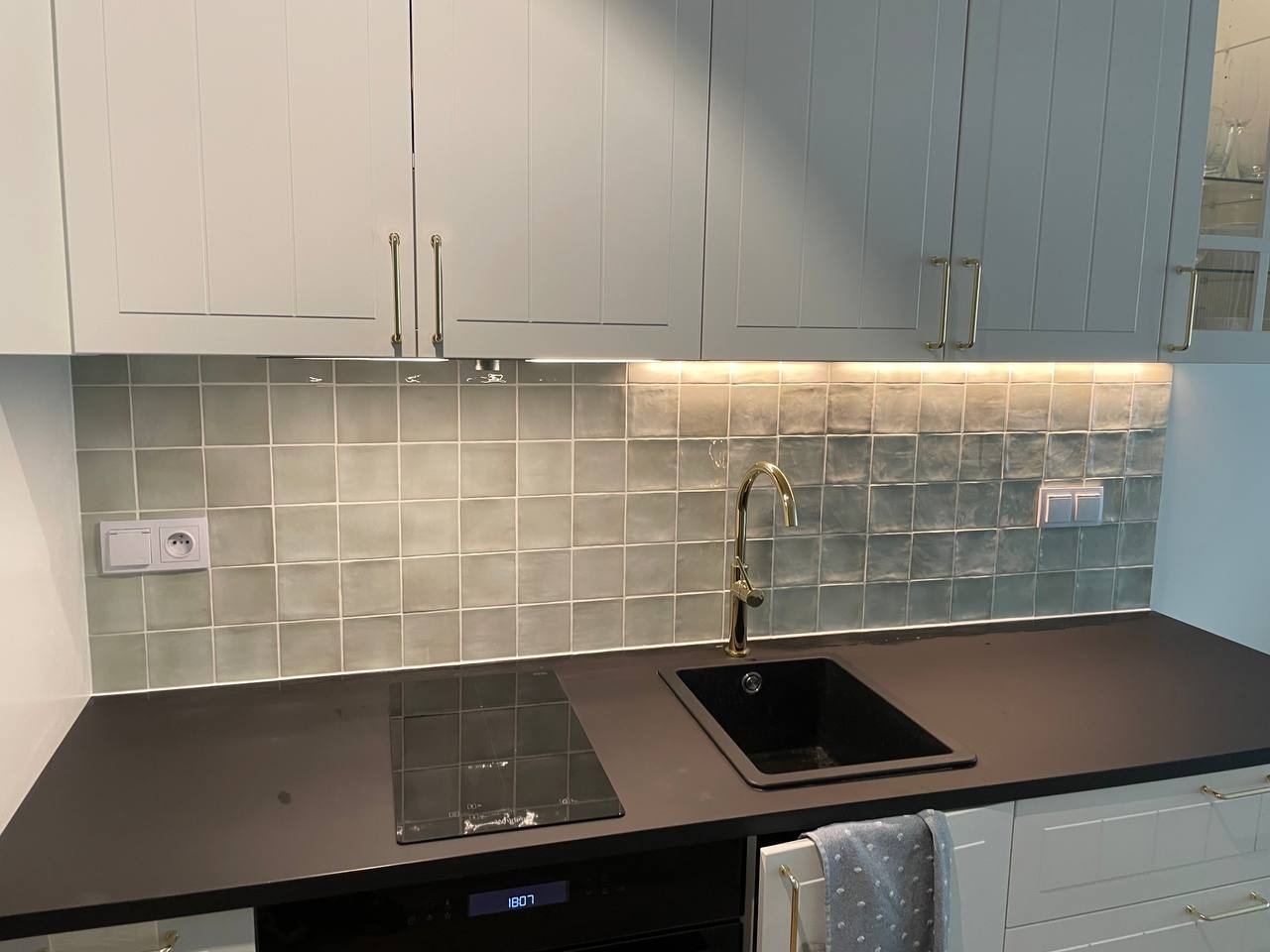 Wykończona kuchnia z szarymi szafkami, złotymi uchwytami i baterią, oraz mozaiką z kwadratowych płytek w odcieniach szarości i zieleni, podświetlonych listwą LED.