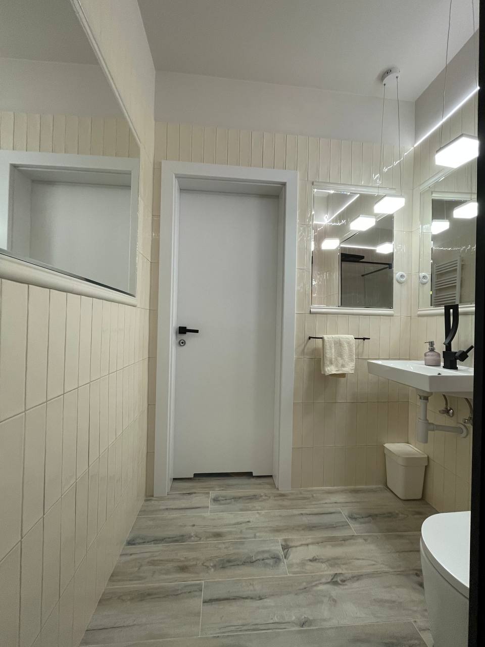 Jasna łazienka z pionowo ułożonymi, kremowymi płytkami na ścianach, imitacją drewna na podłodze, białymi drzwiami i umywalką z czarną armaturą.