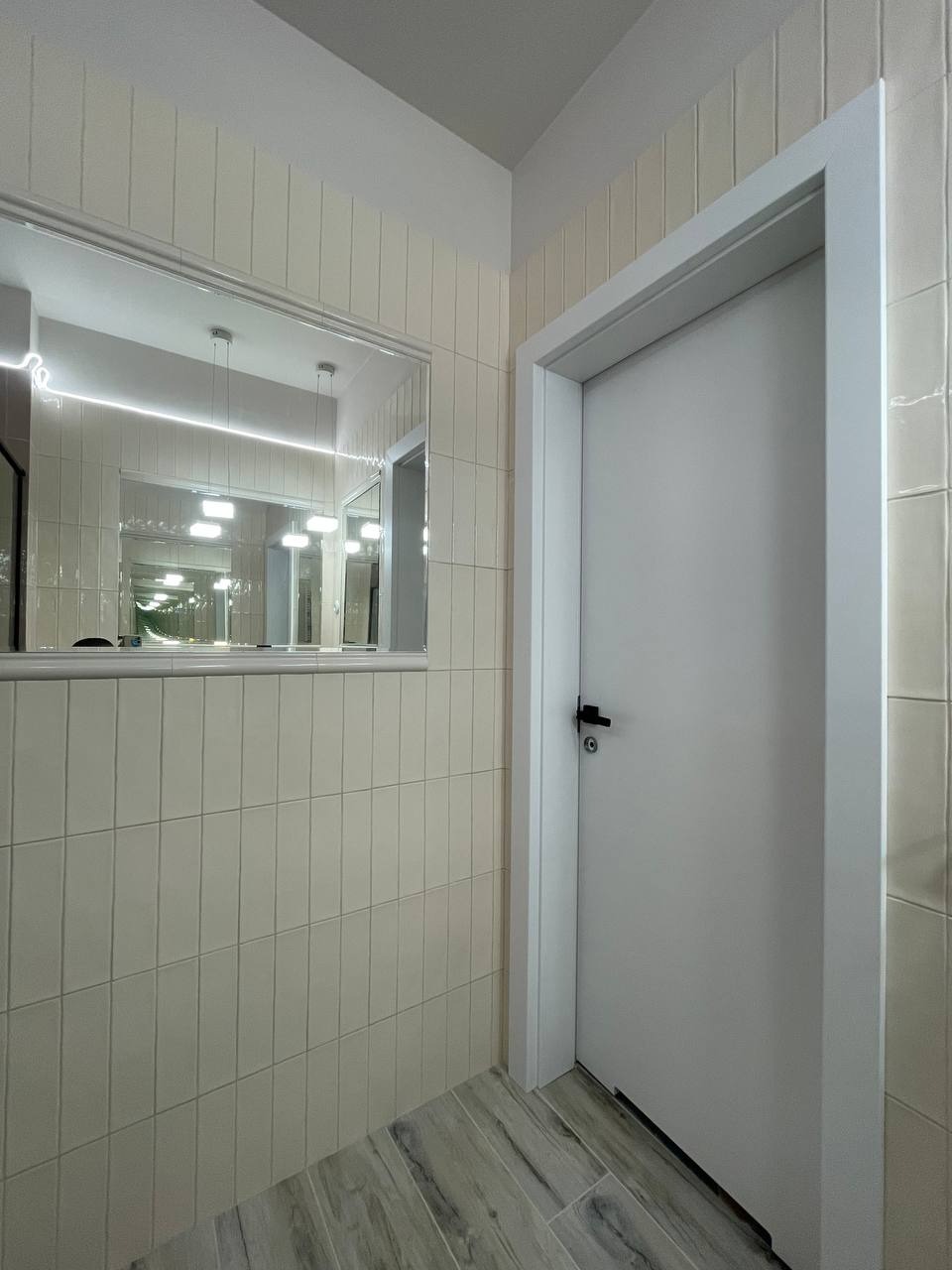 Jasne wnętrze: pionowo ułożone, kremowe płytki na ścianie, lustro z odbiciem oświetlenia i białe drzwi z czarną klamką.