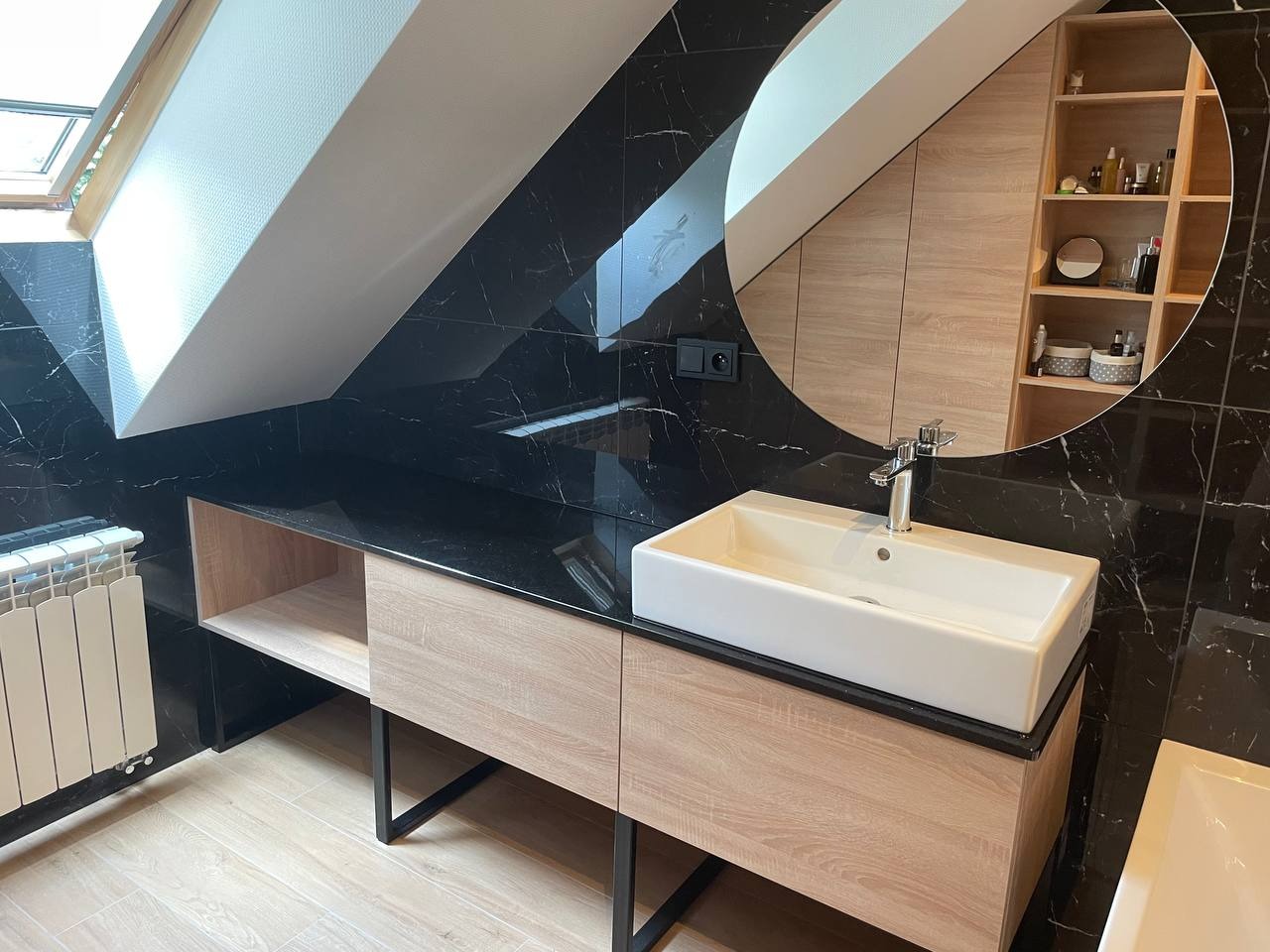 Łazienka na poddaszu z czarnymi marmurowymi płytkami, owalnym lustrem, białą umywalką nablatową na szafce z drewnopodobnym fornirem i grzejnikiem żeberkowym.