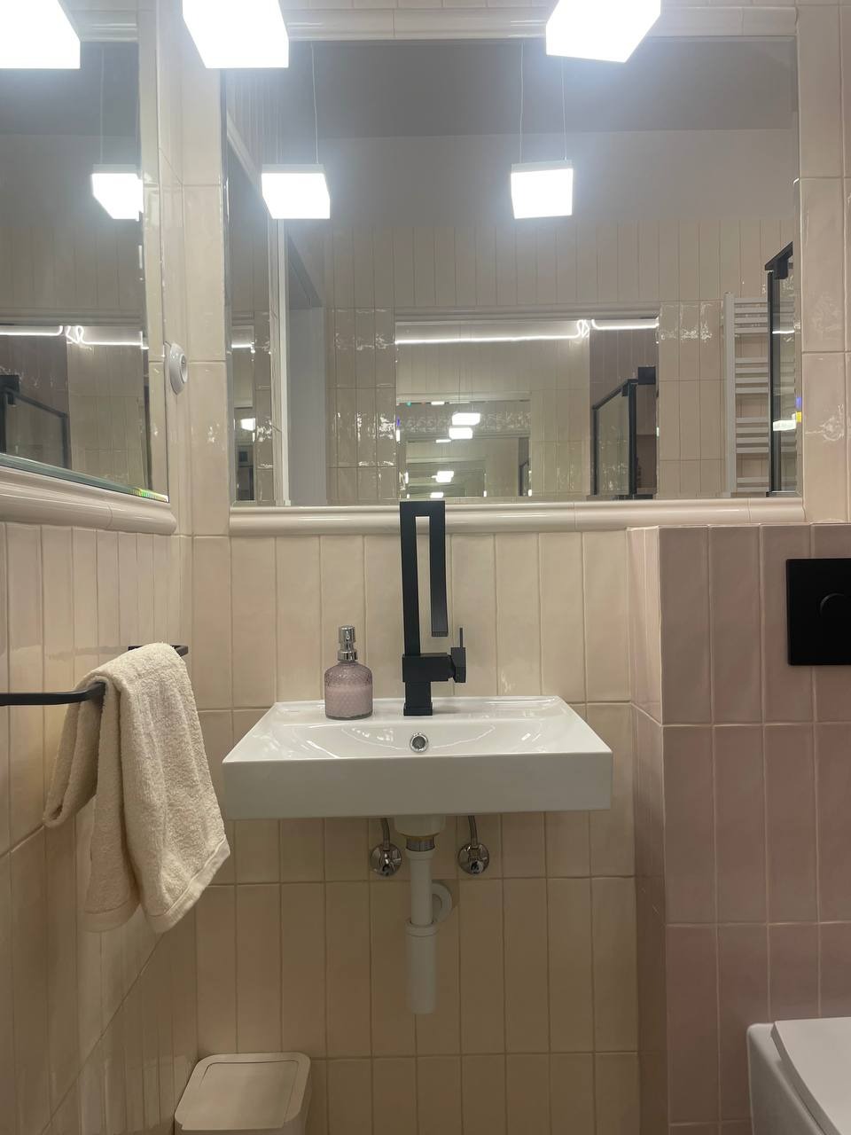 Nowoczesna umywalka nablatowa z czarną armaturą i dekoracyjną butelką na tle beżowych, pionowo ułożonych płytek w łazience z lustrem i kwadratowymi lampami.